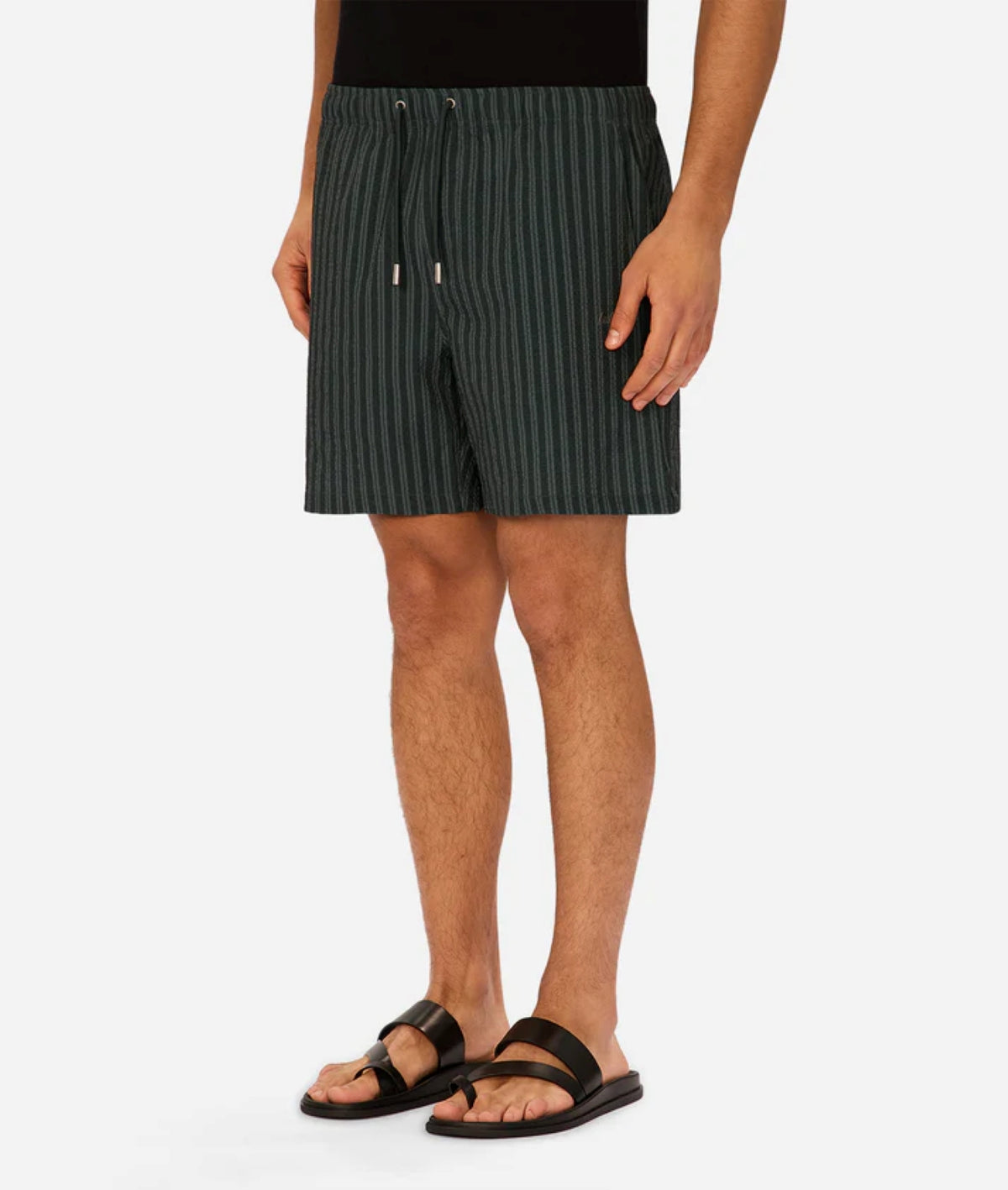 A person is seen from the shoulders down wearing Industrie’s Berkeley Swim Short in dark green seersucker pinstripes, paired with a black shirt and black sandals, standing against a plain white background.