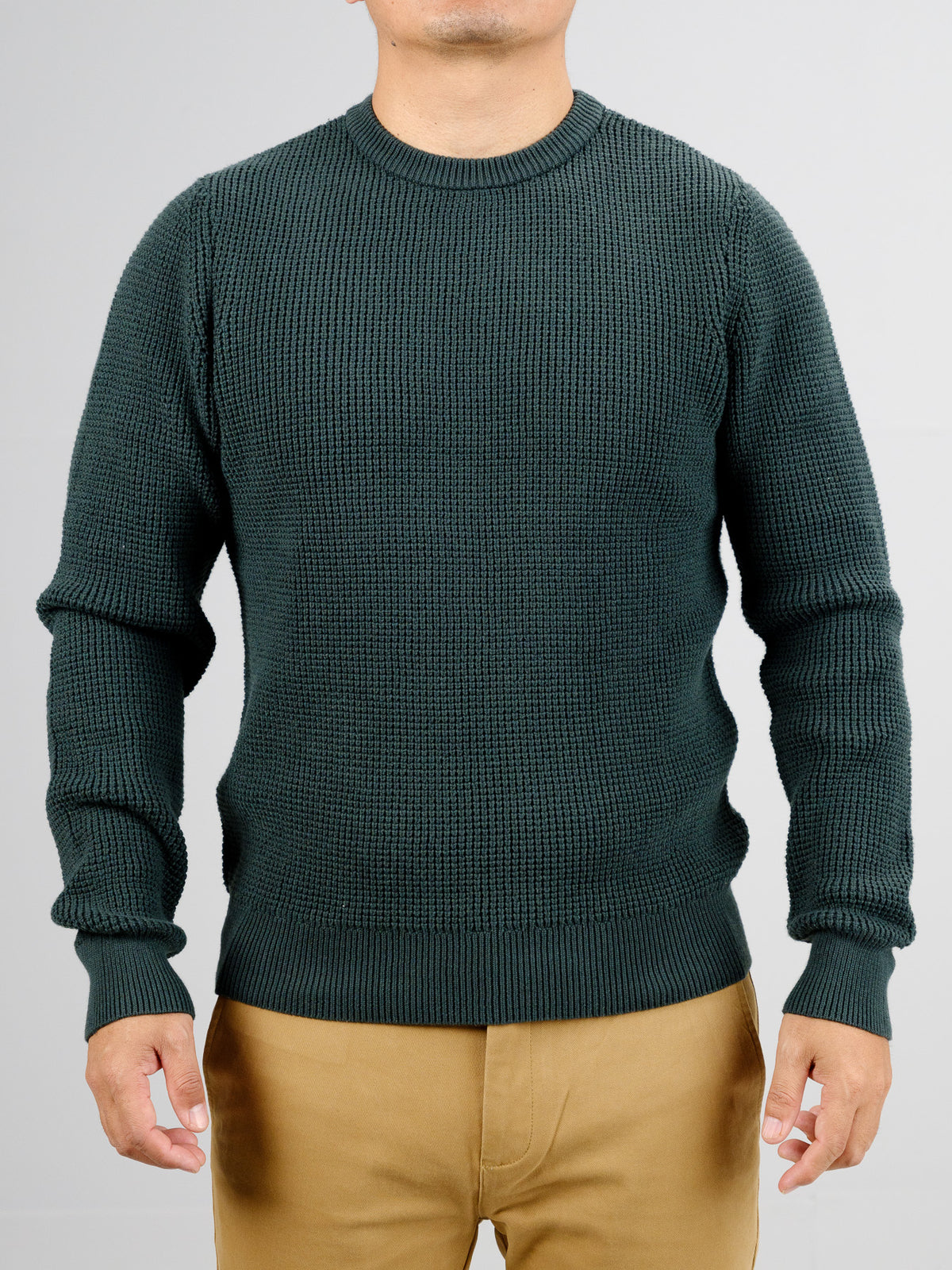 A person in a dark green ID Menswear Ragnar Waffle Knit Sweater, crafted from 100% cotton with long sleeves, stands against a gray background, paired with beige pants. Their face is hidden.