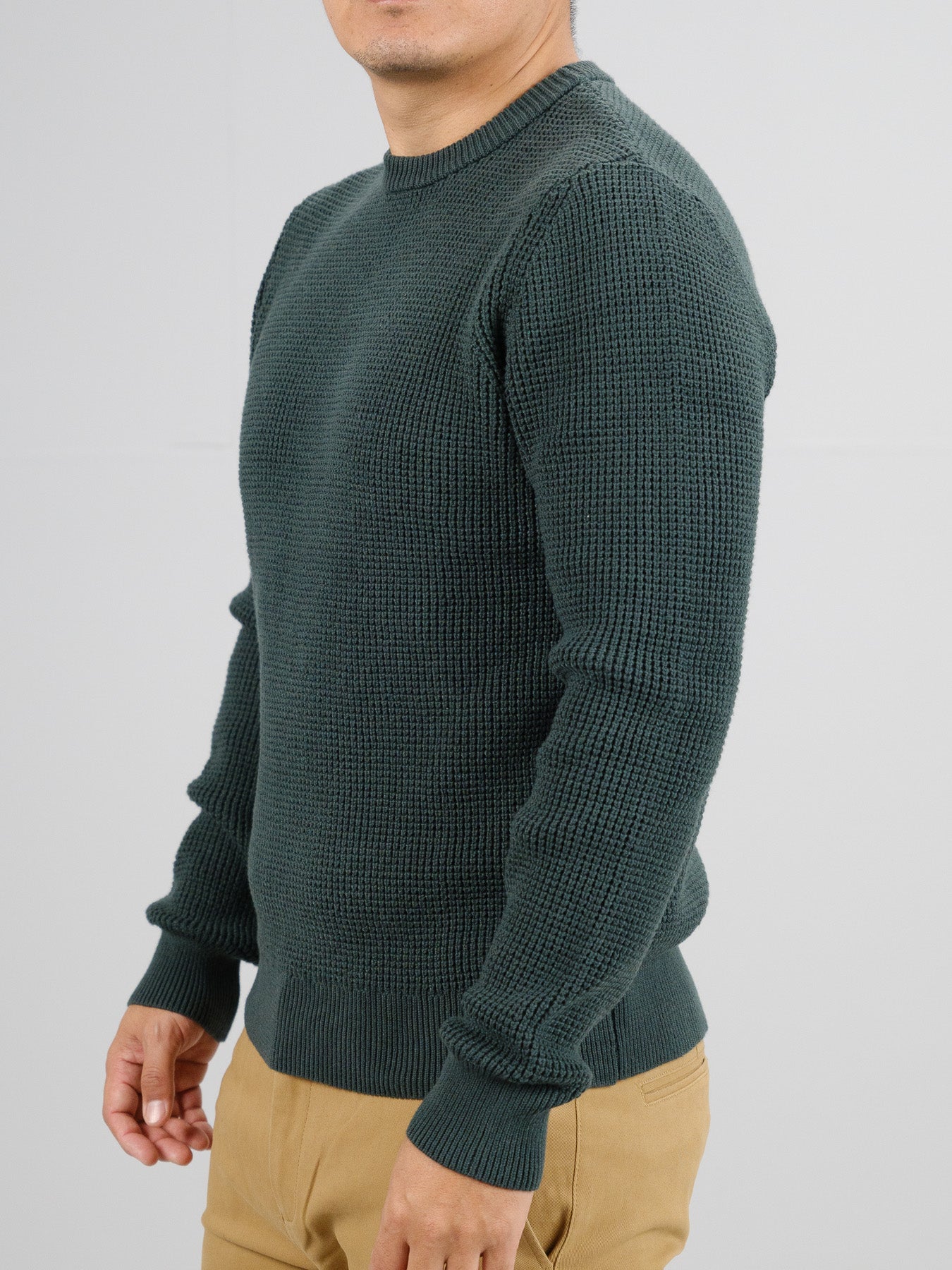 Someone in a textured dark green Ragnar Waffle Knit Sweater by ID Menswear, crafted from 100% cotton, and wearing beige pants stands against a light gray background, with their head out of the frame.