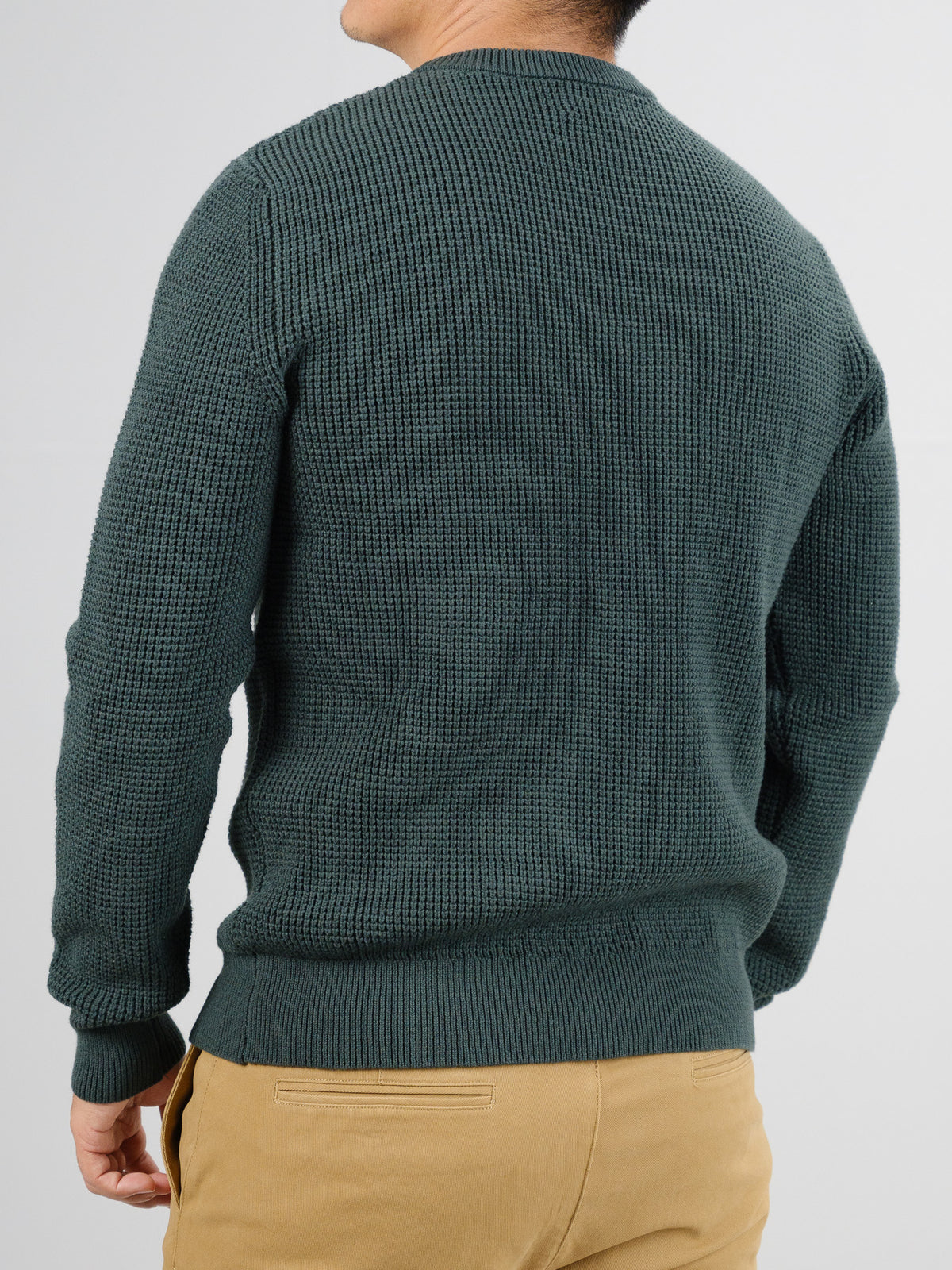 An individual wearing the ID Menswear Ragnar Waffle Knit Sweater, crafted from 100% cotton in dark green, faces away. The textured sweater pairs perfectly with beige pants against a plain white backdrop.