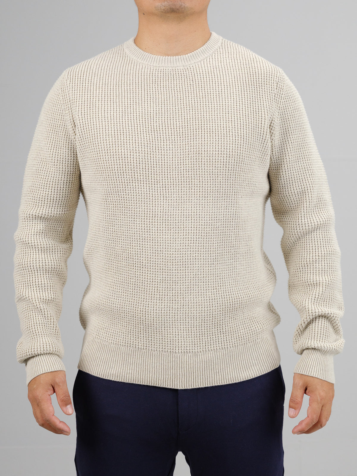 A person wears a beige Ragnar Waffle Knit Sweater by ID Menswear, crafted from 100% cotton with a waffle texture, paired with navy blue pants against a plain gray background. The sweater features a crew neck and long sleeves, displaying only the torso and arms.