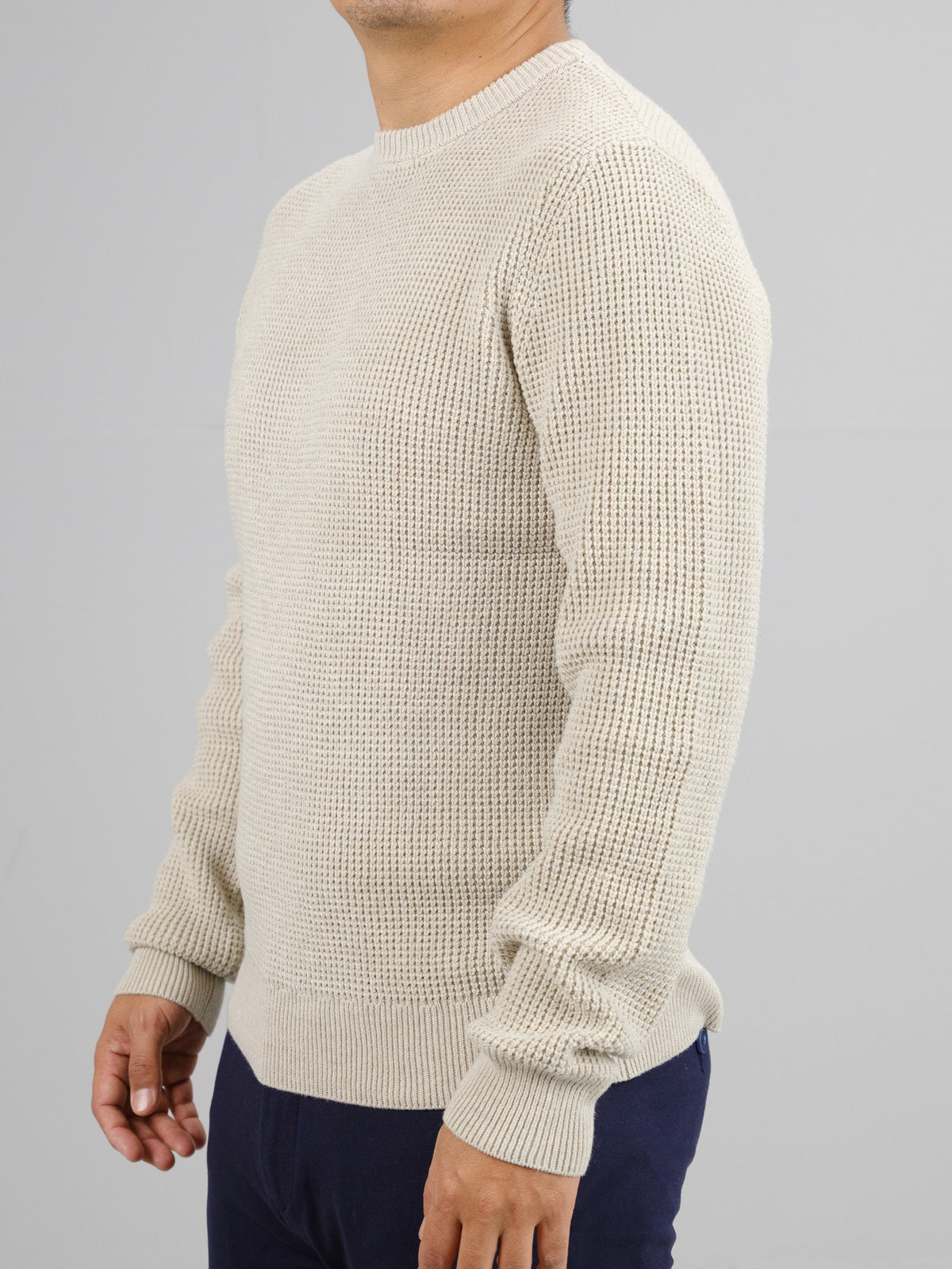 A person wearing the Ragnar Waffle Knit Sweater by ID Menswear in beige, along with navy pants, stands against a plain gray background with their body slightly turned to the side.
