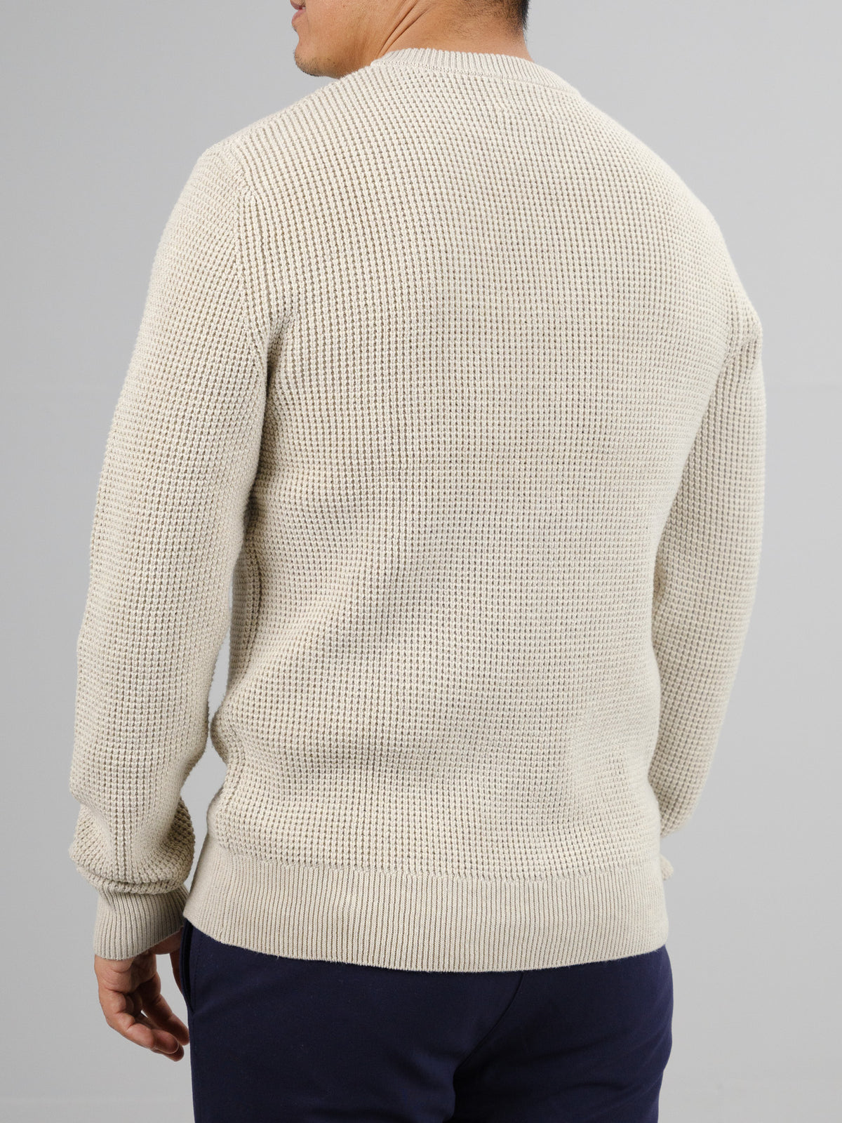 A person in an ID Menswear Ragnar Waffle Knit Sweater, featuring a textured cream design, and dark pants is shown from the back against a plain gray background.