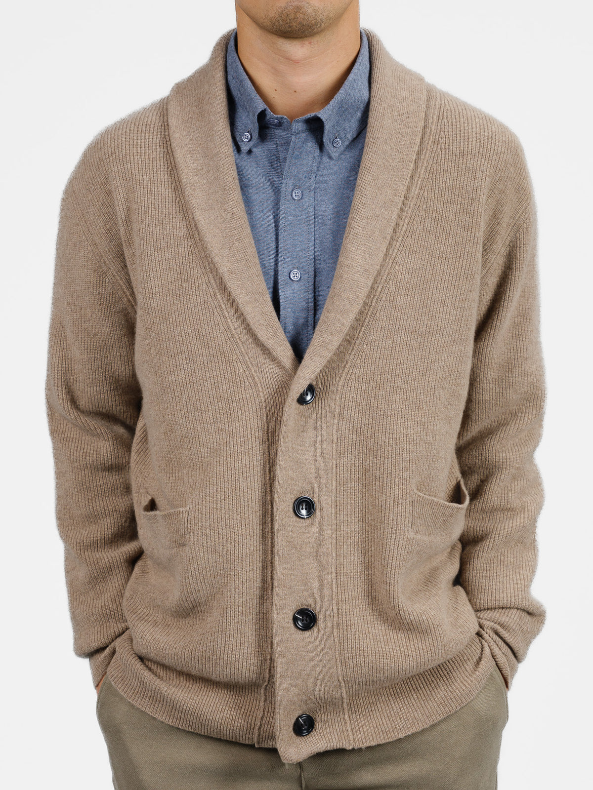 Wearing ID Menswears Basecamp Cashmere Merino Cardigan in beige, with black buttons and front pockets, a person pairs it with a blue button-up and olive pants against a white background—a luxurious winter wardrobe essential.