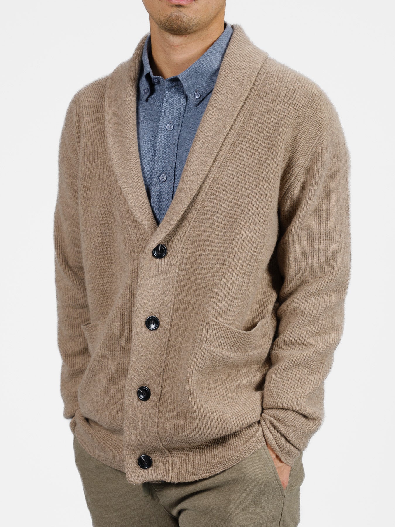 Against a white background, a person wears the ID Menswear Basecamp Cashmere Merino Cardigan, featuring black buttons, a shawl collar, and two pockets. This cozy addition to any winter wardrobe is worn over a blue collared shirt with khaki pants.