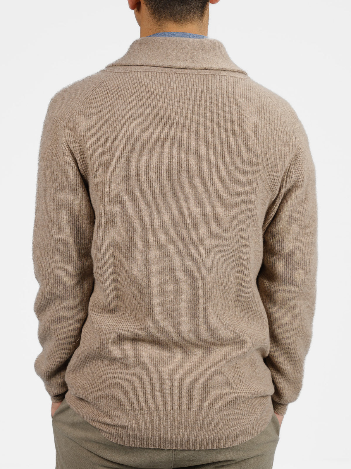 A person is seen from the back wearing ID Menswears Basecamp Cashmere Merino Cardigan in light brown, ribbed knit over a gray shirt and beige pants. Their hands are in their pockets against a white background, highlighting a chic winter wardrobe addition.
