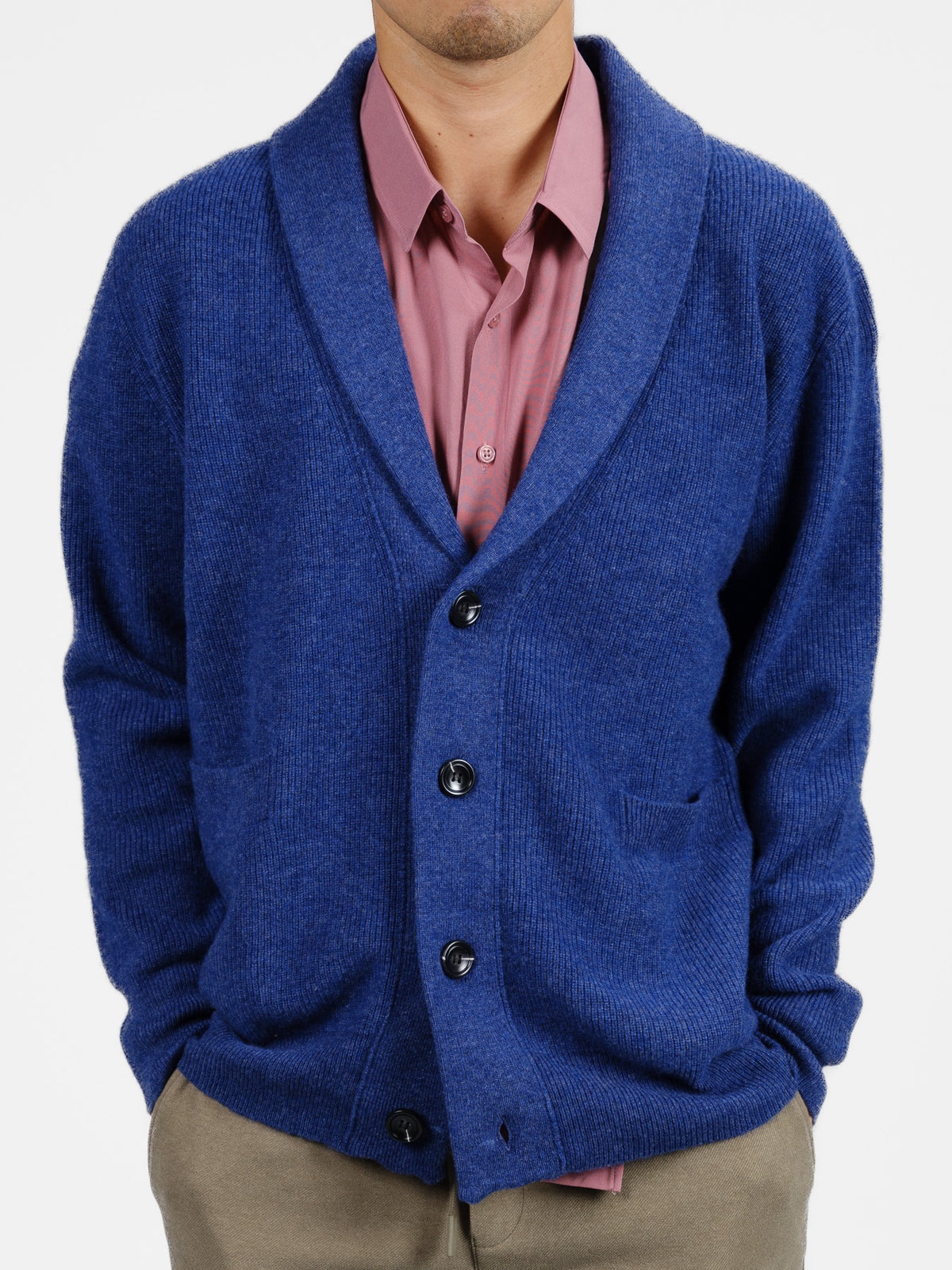 In a white backdrop, someone sports the Basecamp Cashmere Merino Cardigan by ID Menswear, featuring black buttons, a shawl collar, and pockets. Its paired with a pink collared shirt and khaki pants—a winter wardrobe essential.