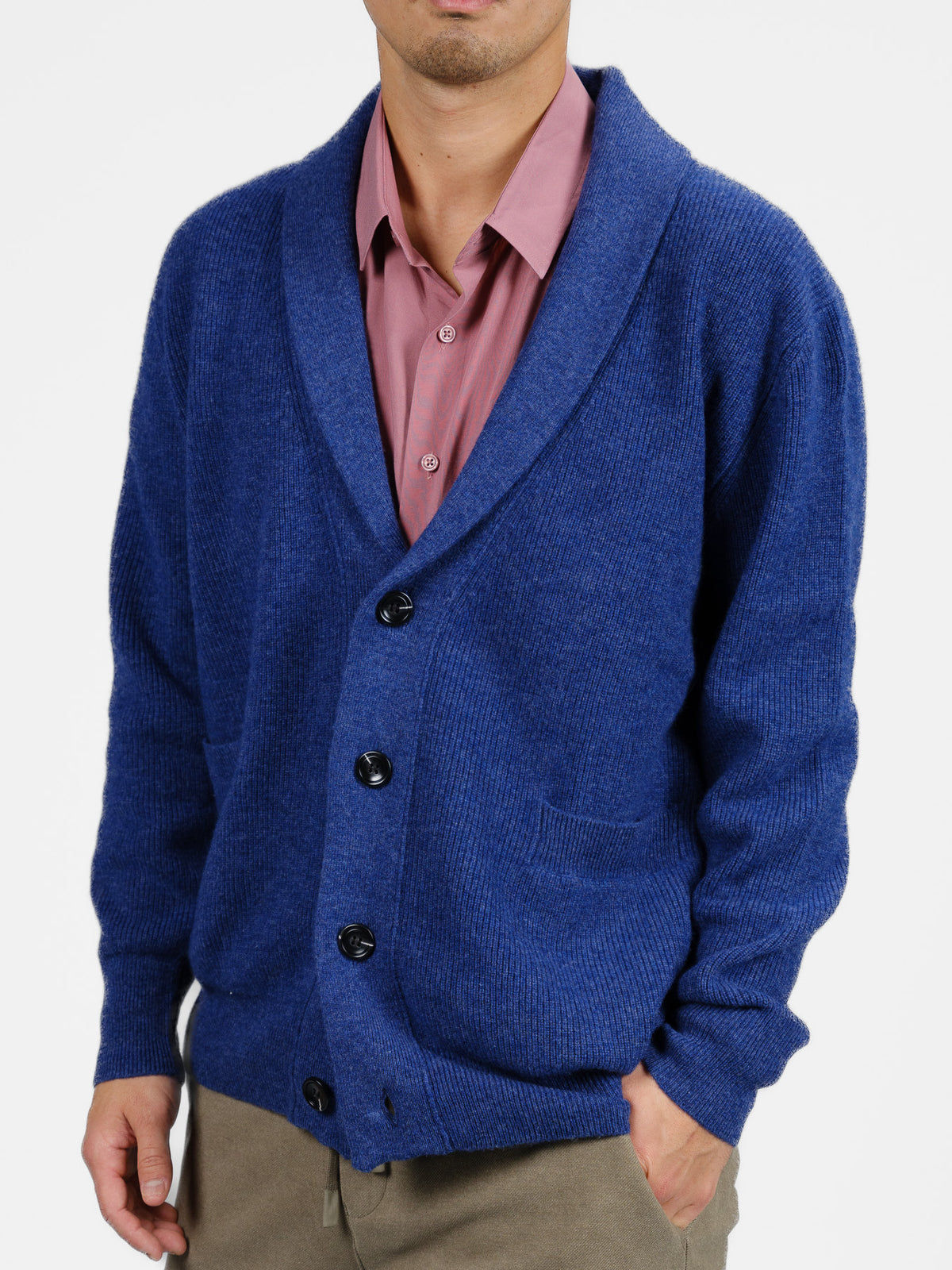 A person embodies cozy elegance in a blue Basecamp Cashmere Merino Cardigan from ID Menswear, layered over a pink shirt with beige pants. The cardigan features a shawl collar, front pockets, and large black buttons, making it perfect for the winter wardrobe against a plain white background.
