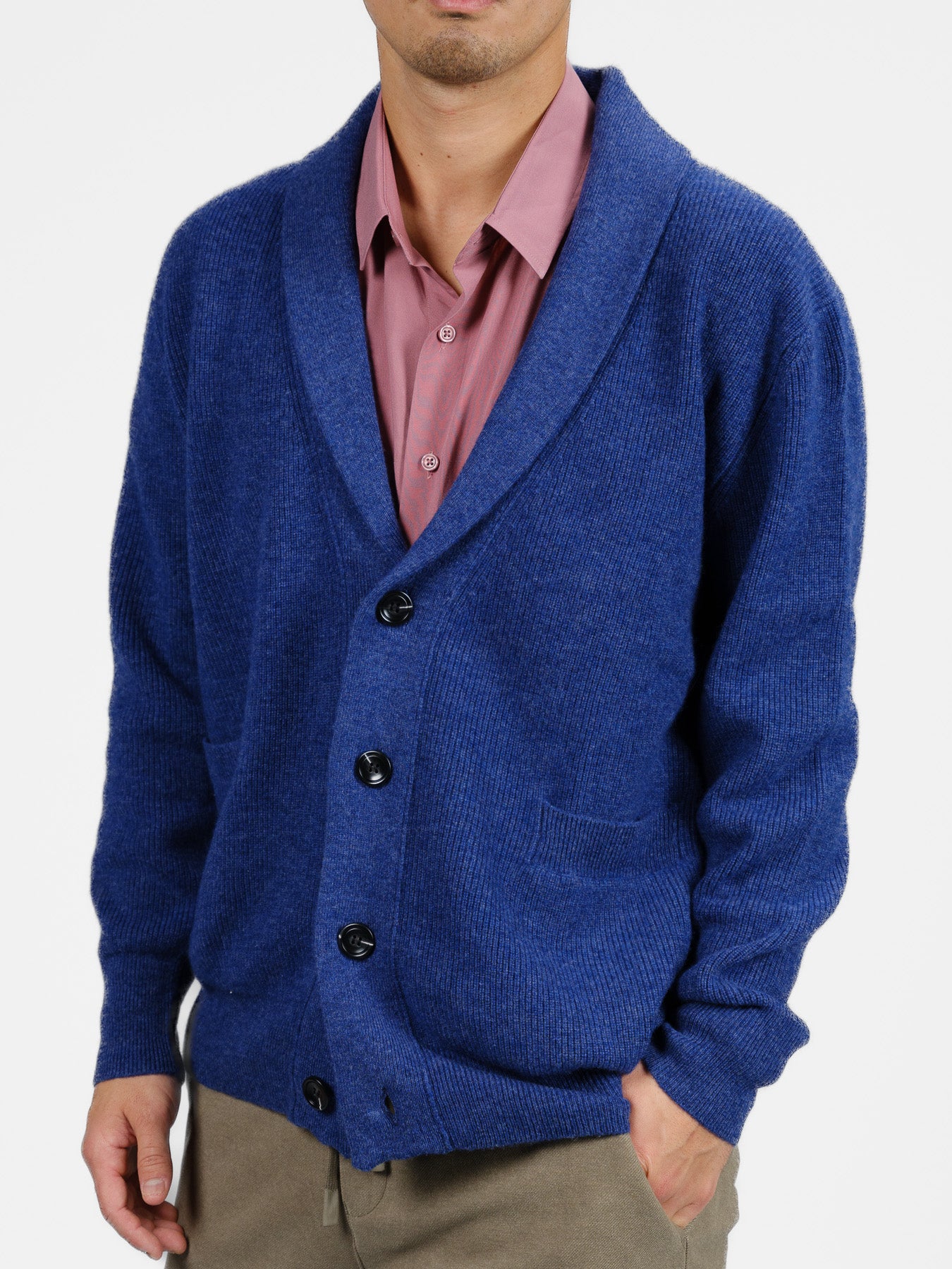 A person embodies cozy elegance in a blue Basecamp Cashmere Merino Cardigan from ID Menswear, layered over a pink shirt with beige pants. The cardigan features a shawl collar, front pockets, and large black buttons, making it perfect for the winter wardrobe against a plain white background.