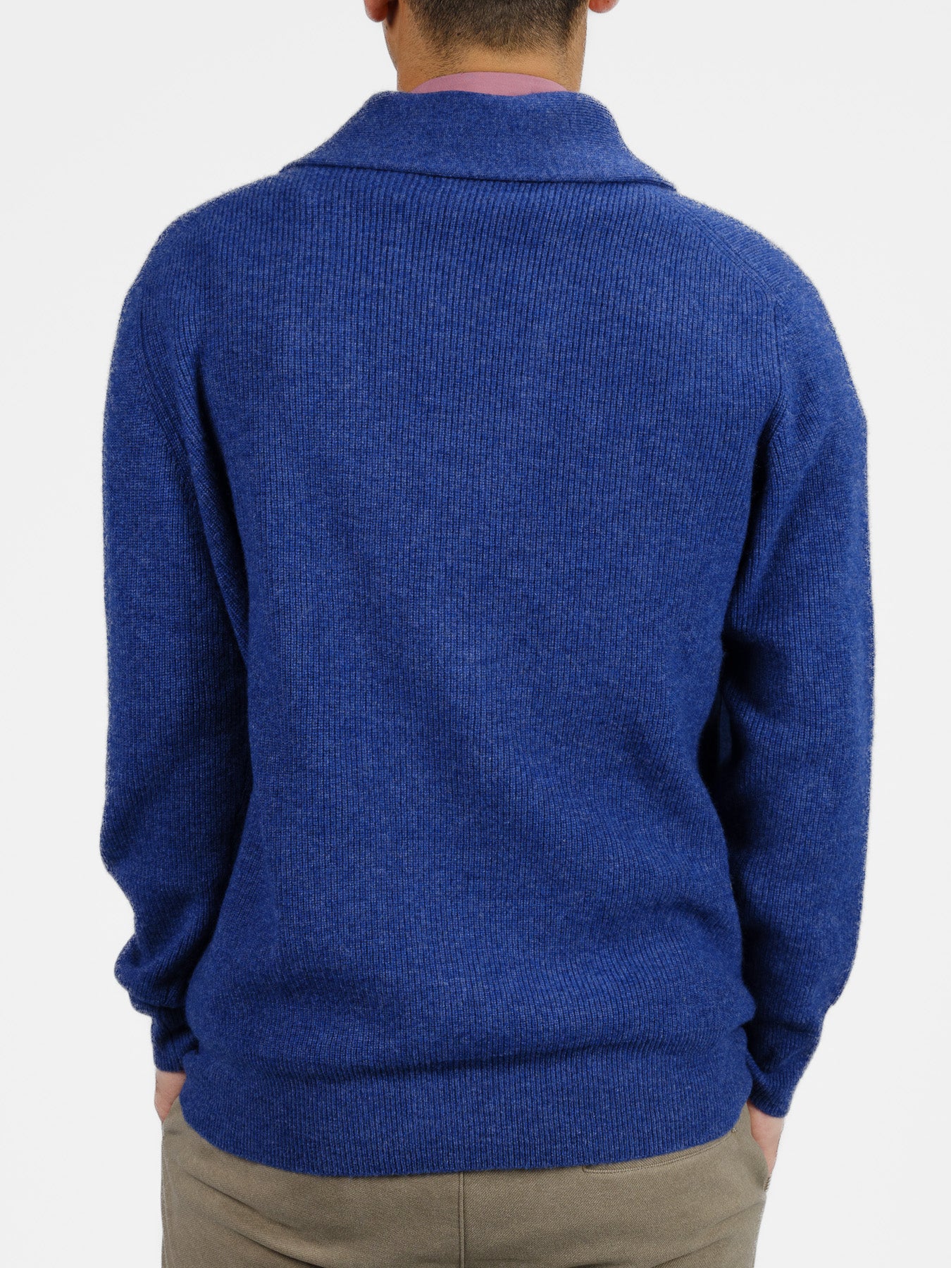 A person wearing ID Menswears Basecamp Cashmere Merino Cardigan in blue, featuring long sleeves and a folded collar, is seen from the back with hands in beige pants pockets. This cozy cardigan is set against a plain white background and is sure to be a winter wardrobe staple.