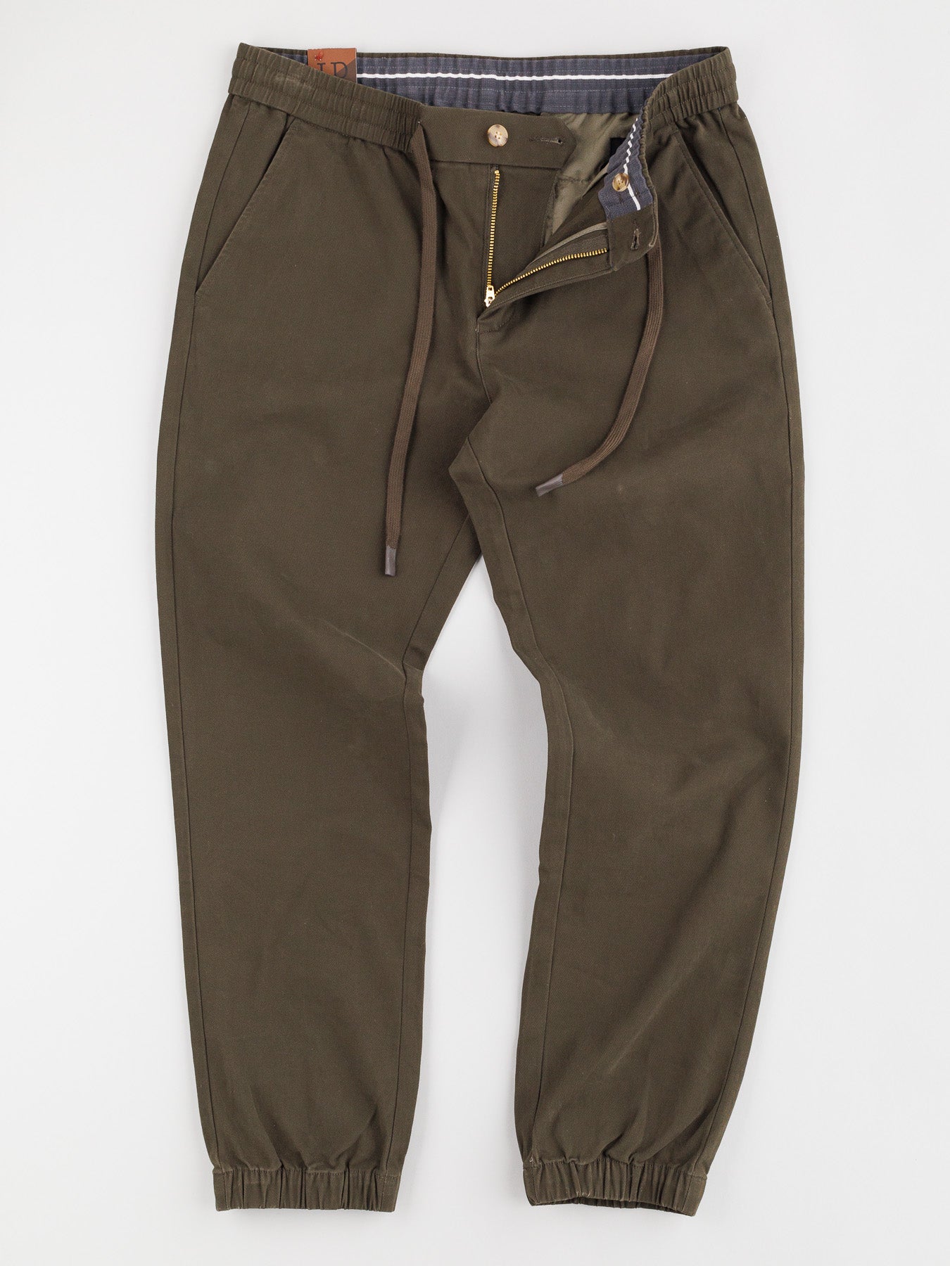 The Dyker Cotton Twill Drawstring Jogger by ID Menswear is made from cotton twill and includes a zipper and button closure. It features olive green elastic cuffs, side pockets, and a chic plaid lining at the waist.