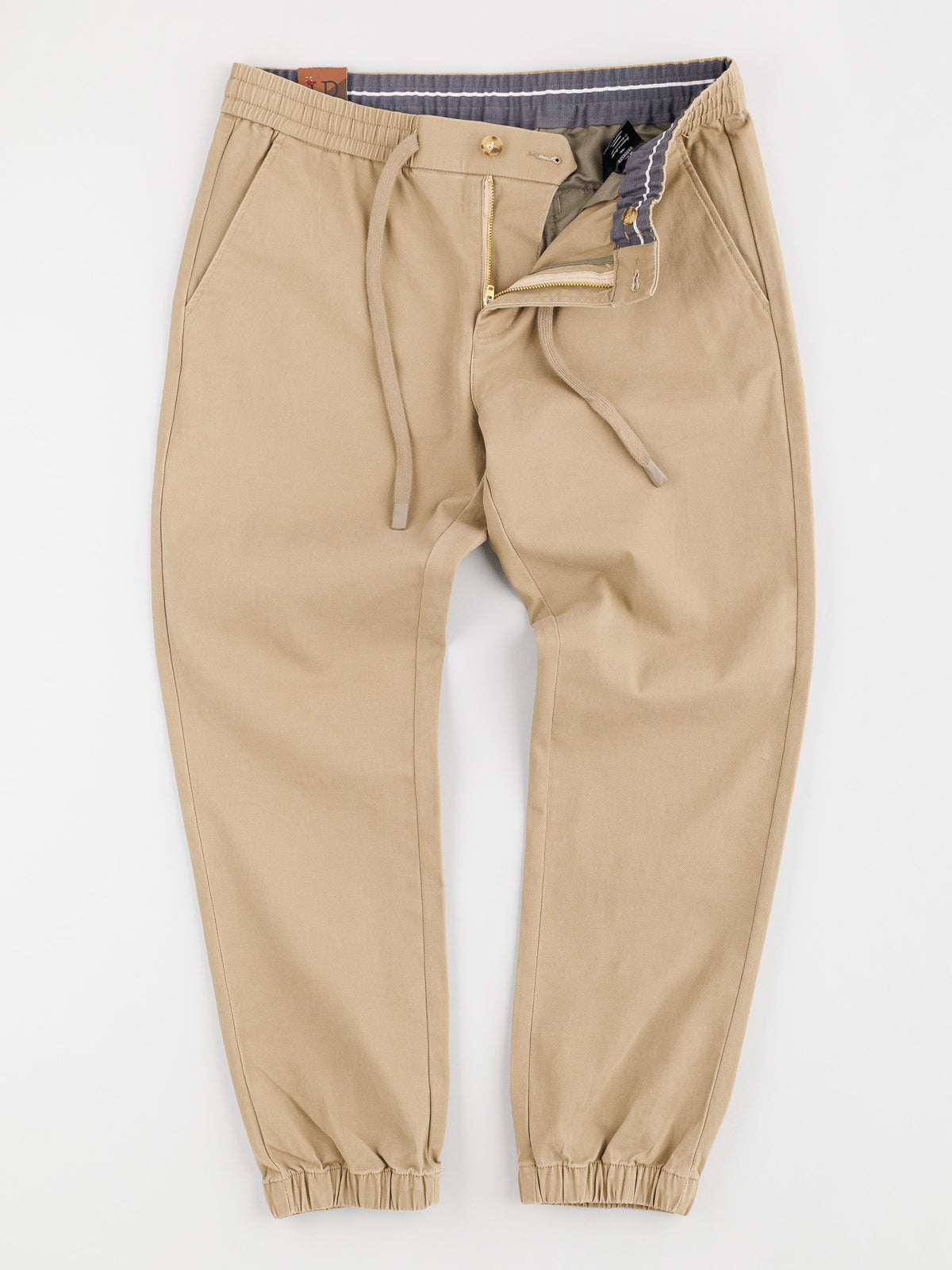 The Dyker Cotton Twill Drawstring Jogger by ID Menswear features beige drawstring joggers with cotton twill, button and zip closure, striped inner waistband, elastic waist cuffs, and side pockets.