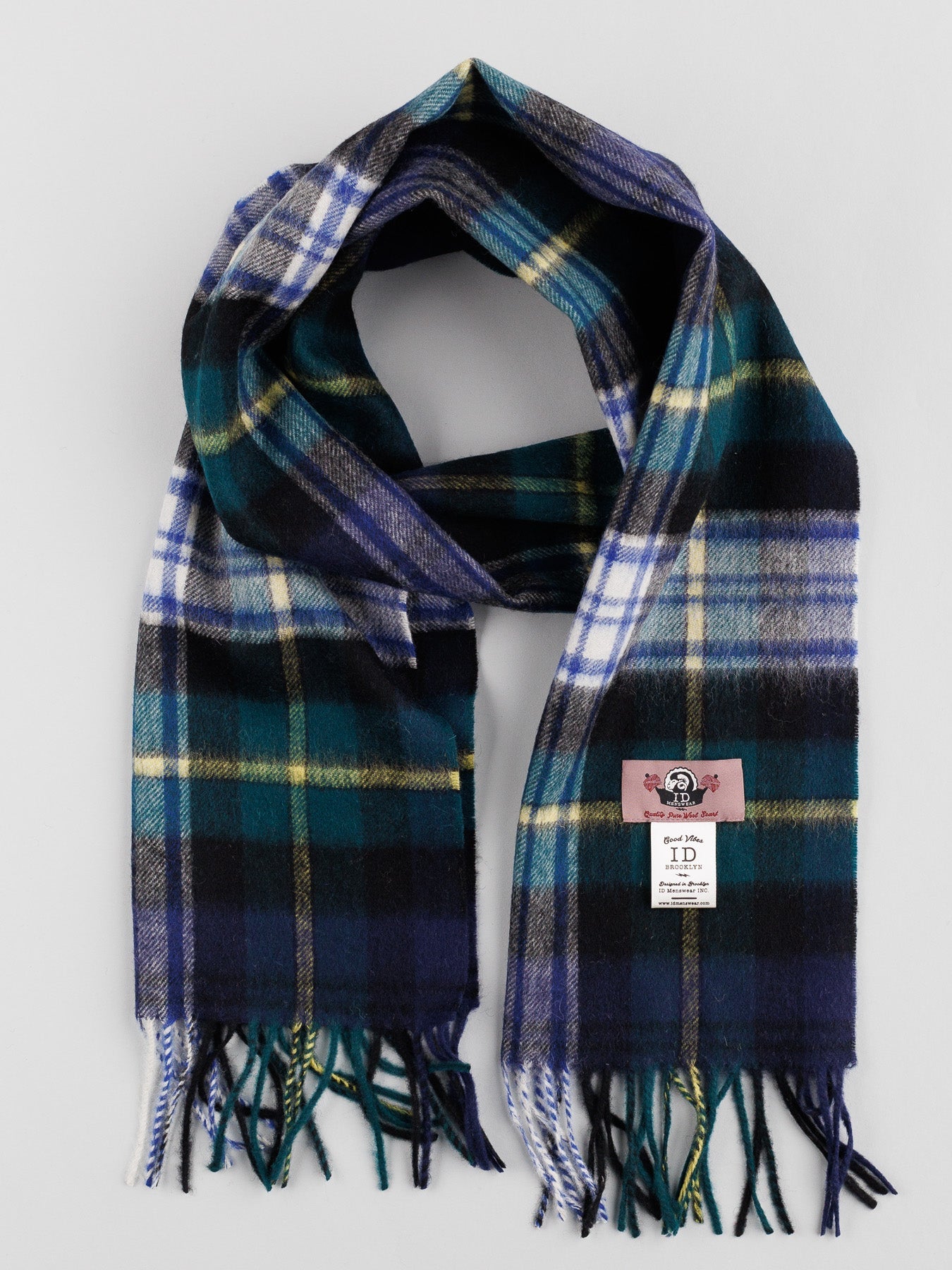 The Wool Plaid Scarf with Self-Fringed Ends by ID Menswear, in blue, green, yellow, and white, is neatly folded. Featuring fringed edges and a label with an illustration of a woman’s face, this soft wool scarf complements any winter wardrobe against a light gray background.