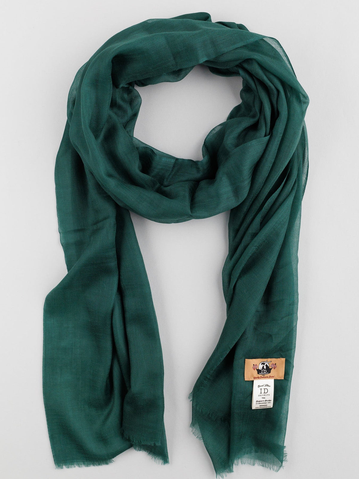Elegantly draped on a light gray background, the Cashmere Air Thin Shawl by ID Menswear is in dark green and made from ultra-soft material with a delicate texture. It includes a small beige tag with a logo and text, making it both stylish and luxurious.