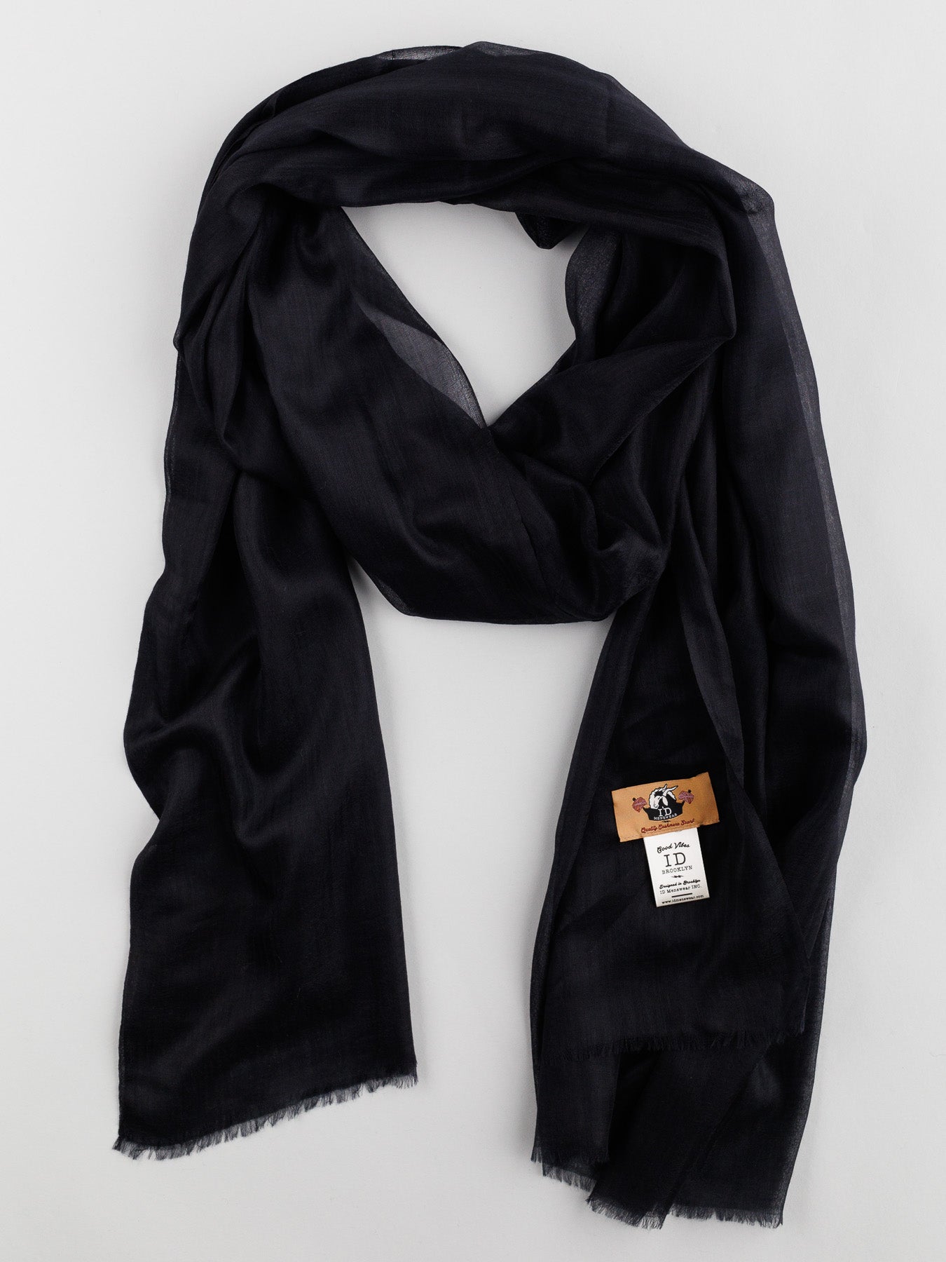 The Cashmere Air Thin Shawl from ID Menswear is elegantly draped, showcasing its ultra-soft 100% cashmere texture. A logo-labeled tag is near the edge, and a slight fringe on the ends enhances its sophisticated appearance.