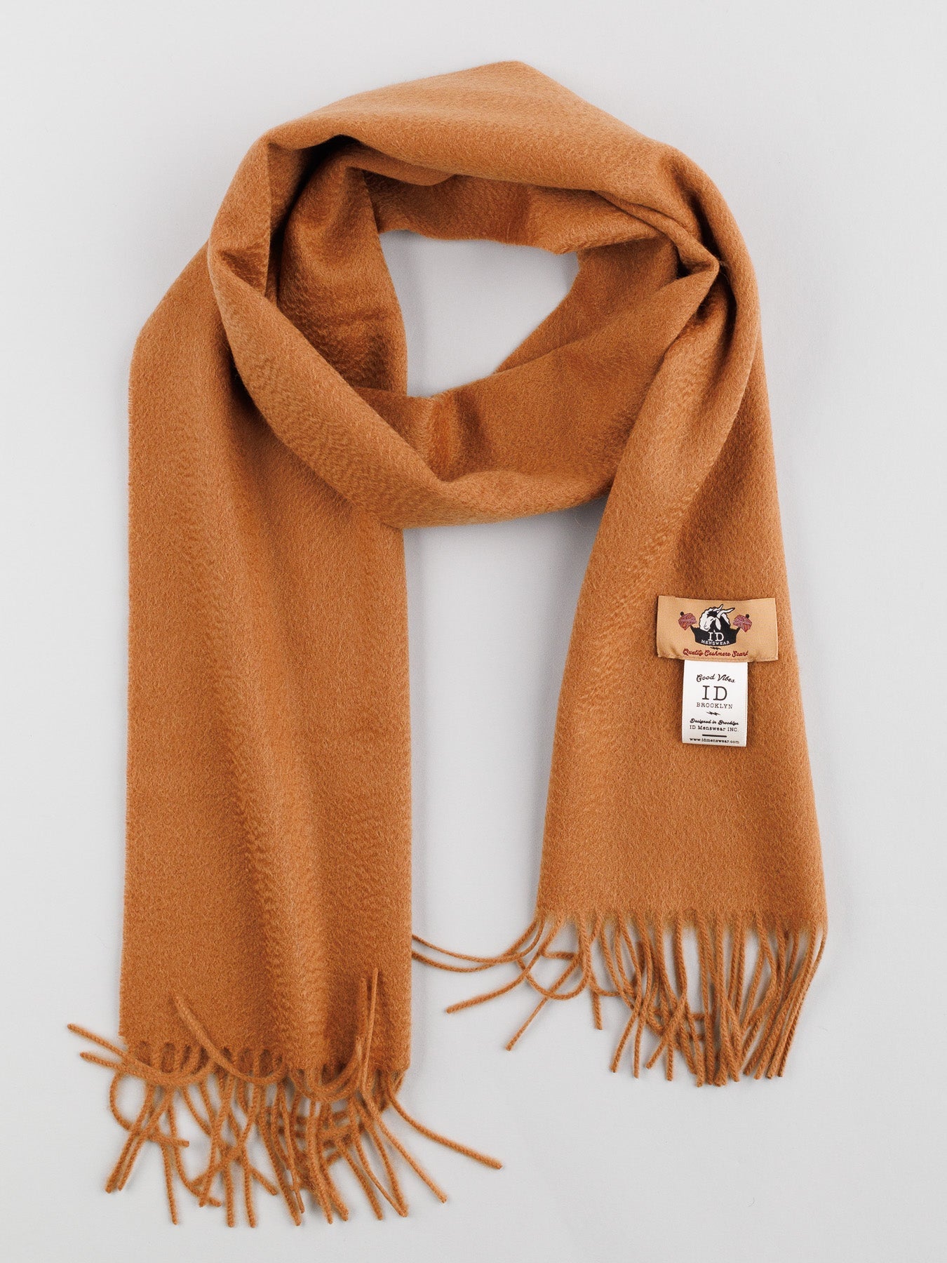 Elegantly folded, the soft Cashmere Scarf with Self-Fringed Ends from ID Menswear features a light brown hue and subtle herringbone texture. A small rectangular label with text and logo adorns it, making this versatile piece a perfect addition to any winter wardrobe.