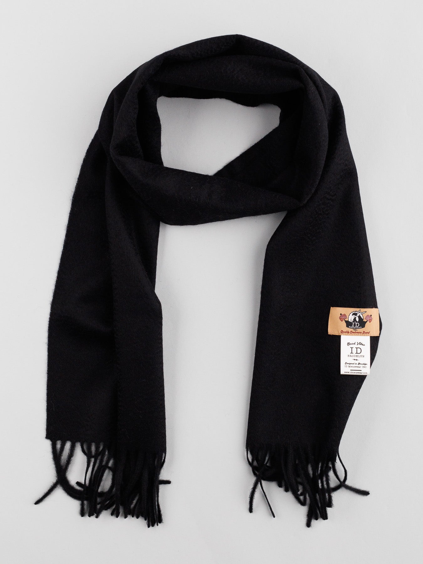 A black ID Menswear Cashmere Scarf with self-fringed ends is displayed on a light gray background, exuding timeless elegance. It sports a small tan tag with printed text and logo—an essential winter wardrobe addition.