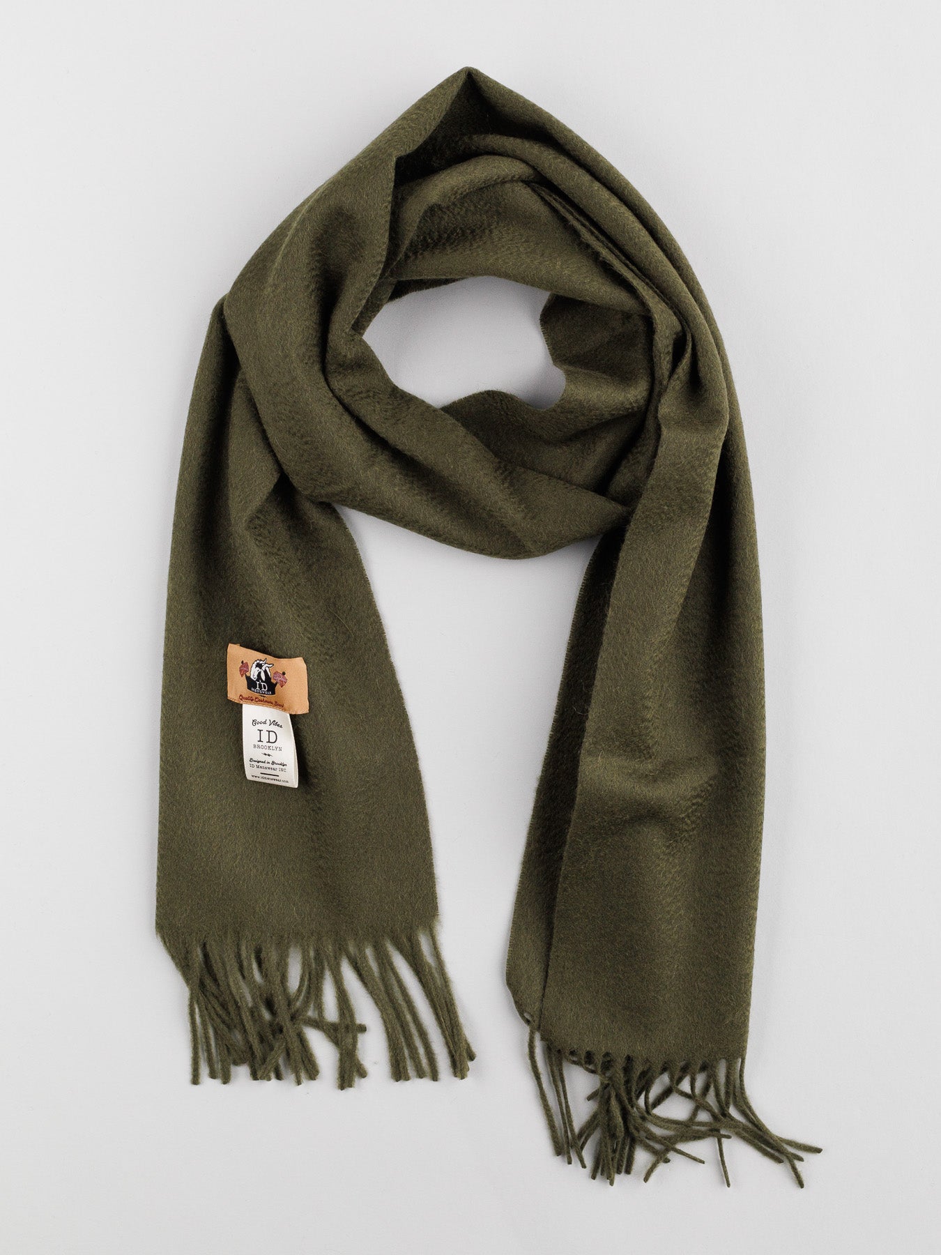 A Cashmere Scarf with Self-Fringed Ends by ID Menswear is displayed flat on a white background, exuding timeless elegance. The dark green scarfs fringed ends and small logo tag enhance this winter wardrobe staples charm.