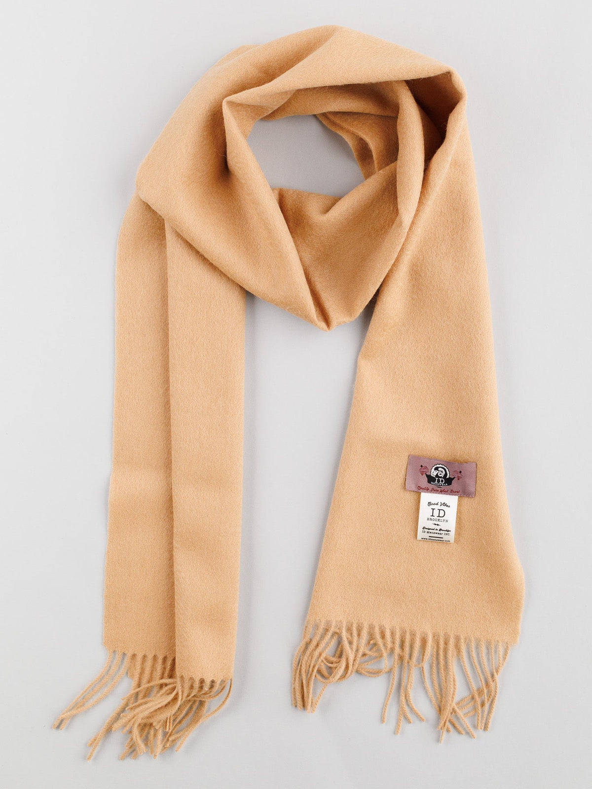 A beige Wool Scarf with Self-Fringed Ends from ID Menswear, laid flat, displays a small pink cartoon tag and a white label showing text and logo beneath it. This stylish winter accessory features an elegantly folded classic loop on top.