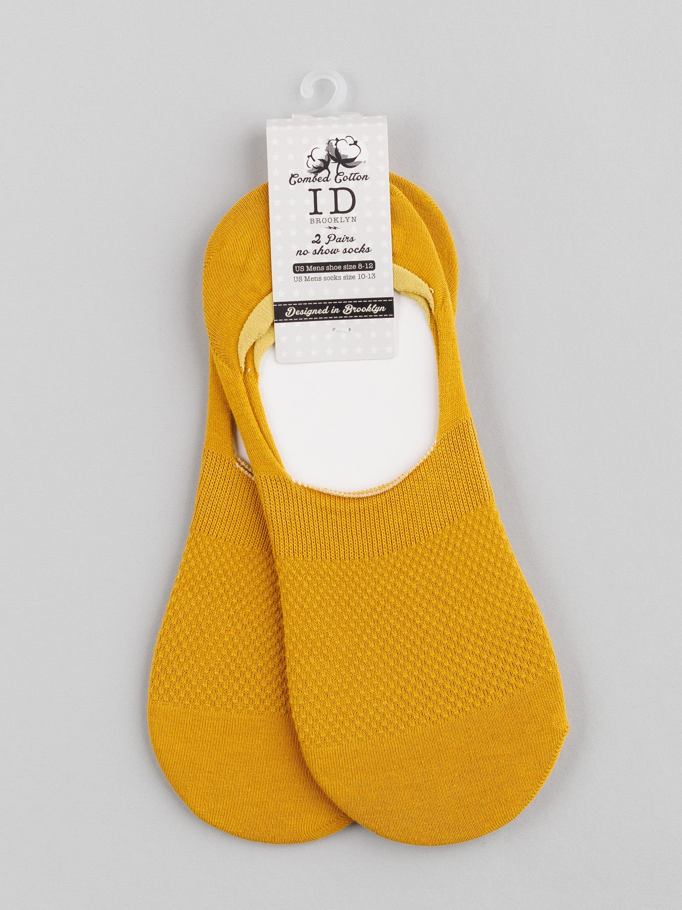 A pair of mustard yellow ID Menswear M1 No Show Socks made from soft combed cotton with a textured pattern are shown on a light gray background, attached to labeled cardboard packaging, ideal for wearing with low-cut shoes.