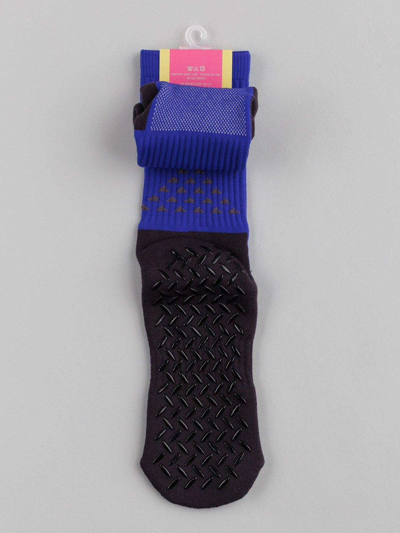 A single Grip Sports Sock by ID Menswear, featuring a blue upper and darker purple foot section, hangs from a hook. It showcases black anti-slip dots on the sole for performance and comfort. The top label is pink with text.