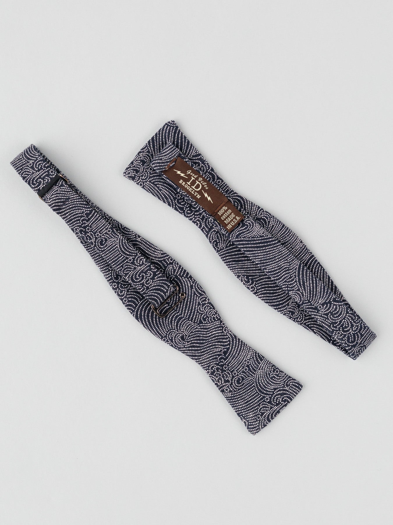 Two Japanese Nami Printed Bow Ties by ID Menswear, handmade in Brooklyn with a bold Japanese wave pattern, are laid flat on a white surface; one displays a brown label with text and sizing.