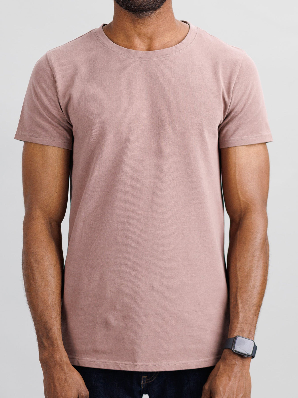 A person in a slim-fit City Organic Cotton Tee by ID Menswear, light brown in color, and a black smartwatch stands against a neutral backdrop with their arms relaxed at their sides.
