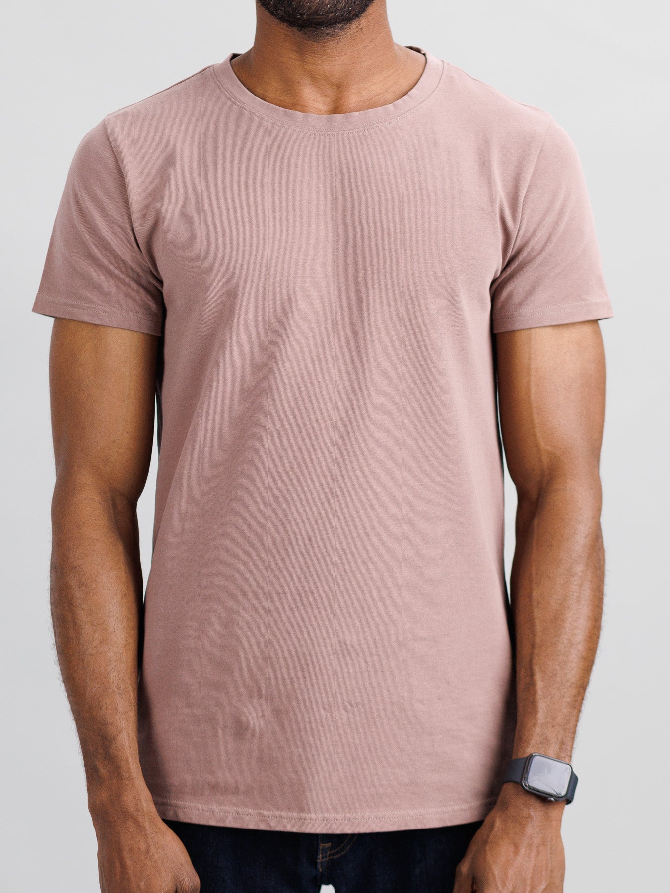 A person in a slim-fit City Organic Cotton Tee by ID Menswear, light brown in color, and a black smartwatch stands against a neutral backdrop with their arms relaxed at their sides.