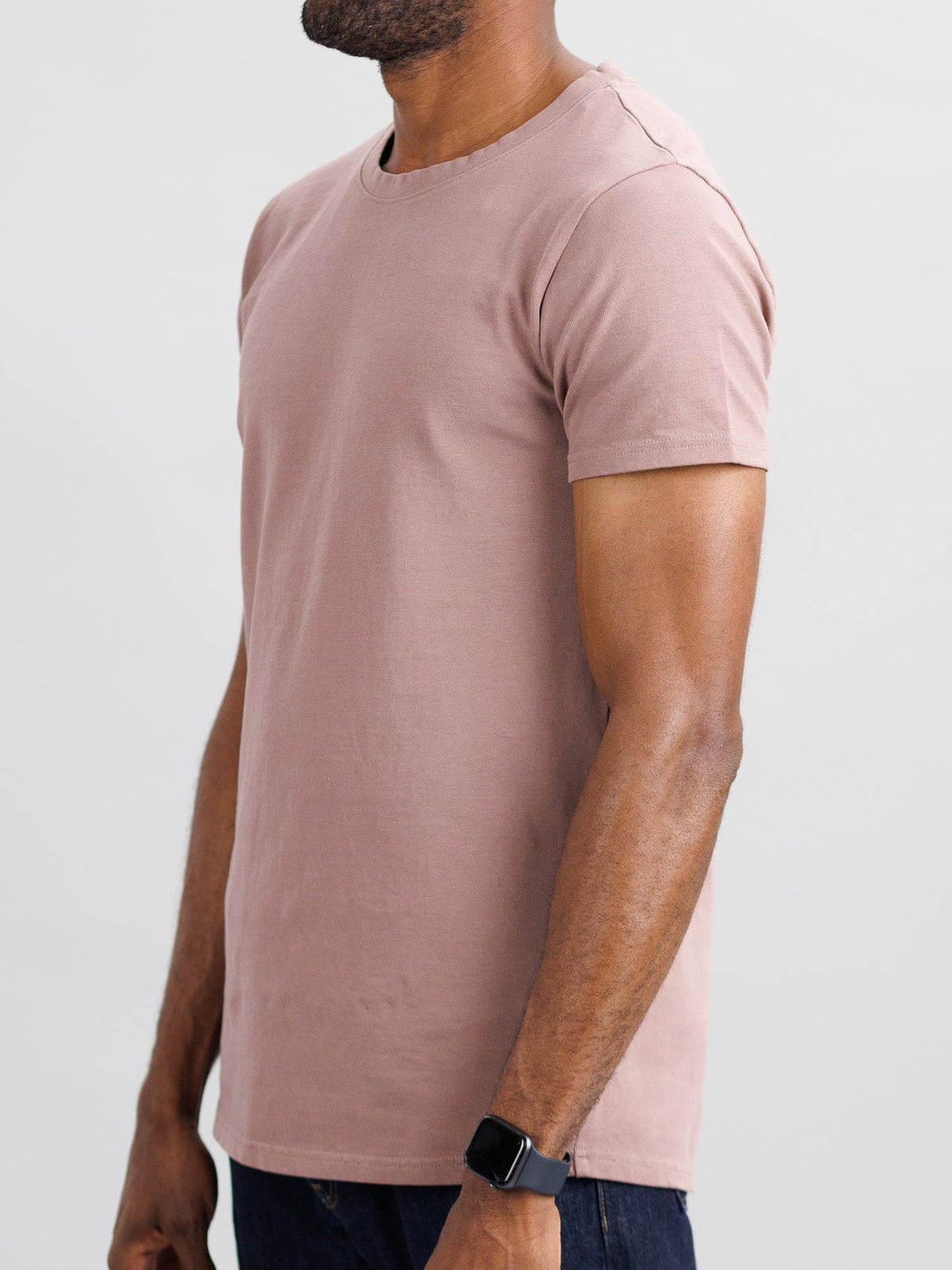 A person sports ID Menswears slim fit City Organic Cotton Tee in pinkish-brown and dark jeans, accessorized with a smartwatch on their left wrist. A light gray background enhances the minimalist vibe in the cropped image above their head.