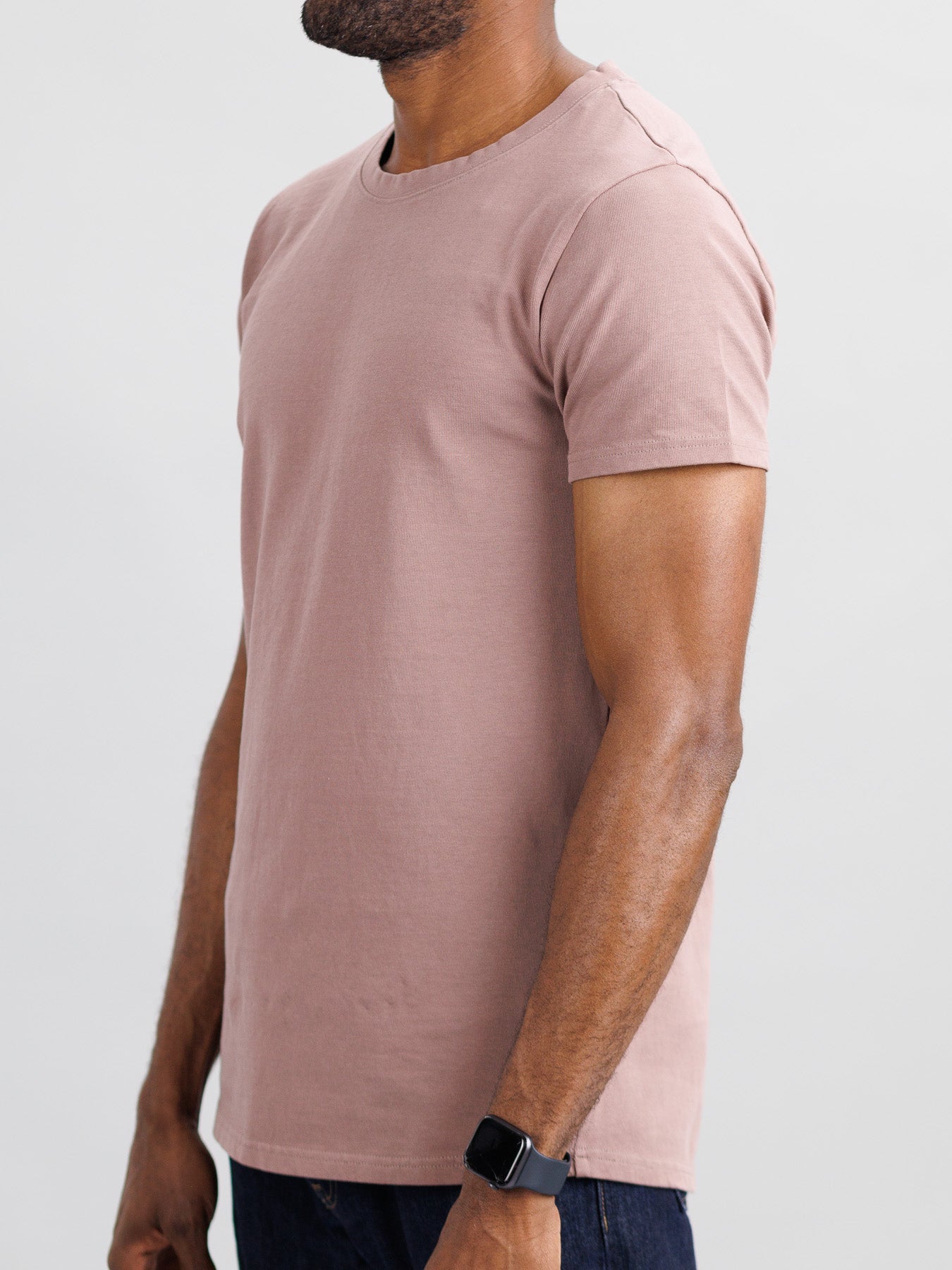 A person sports ID Menswears slim fit City Organic Cotton Tee in pinkish-brown and dark jeans, accessorized with a smartwatch on their left wrist. A light gray background enhances the minimalist vibe in the cropped image above their head.