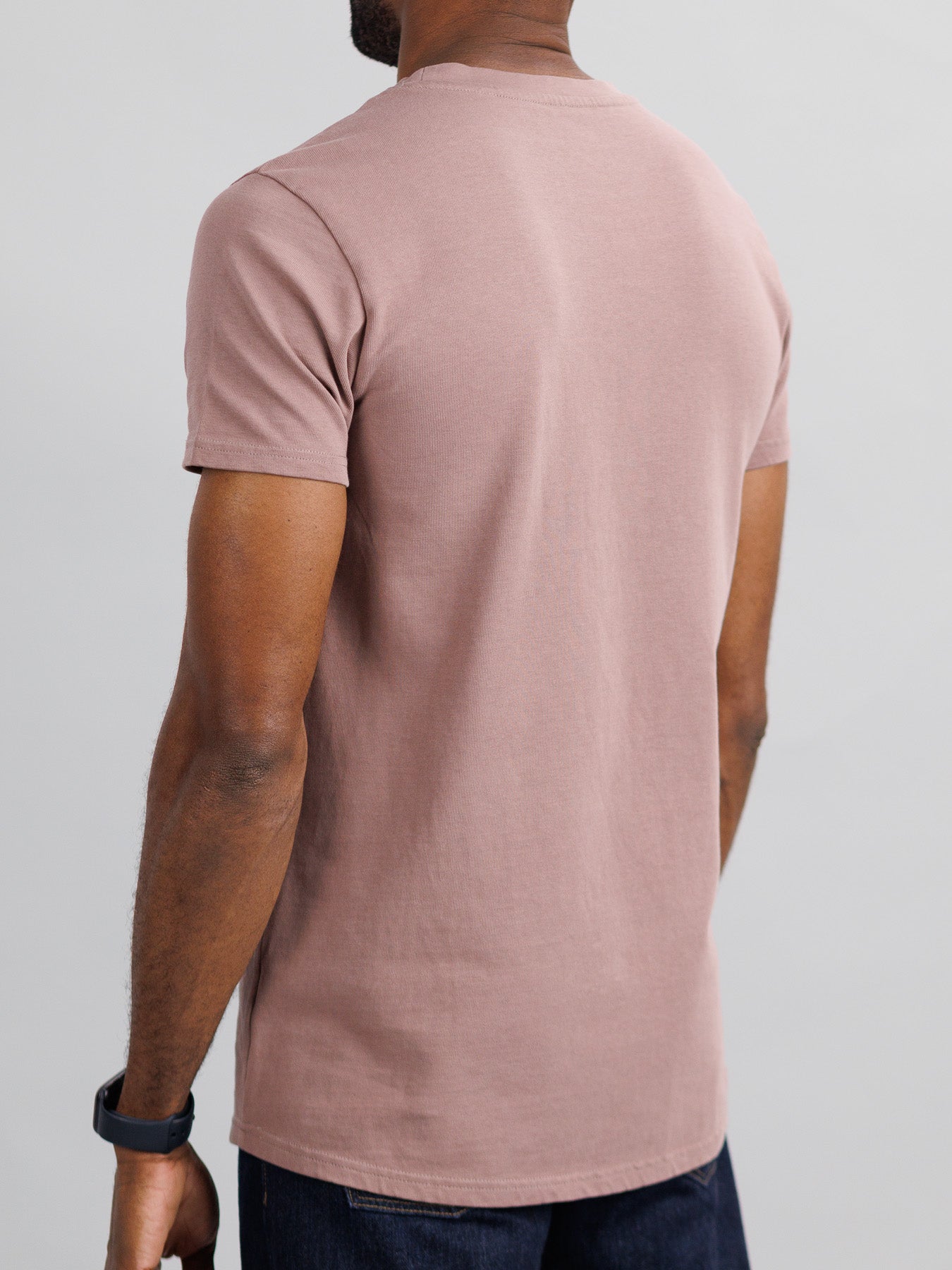 A man wears ID Menswears City Organic Cotton Tee in slim fit mauve, paired with dark jeans. On his left arm is a black smartwatch. He stands against a plain light gray background.