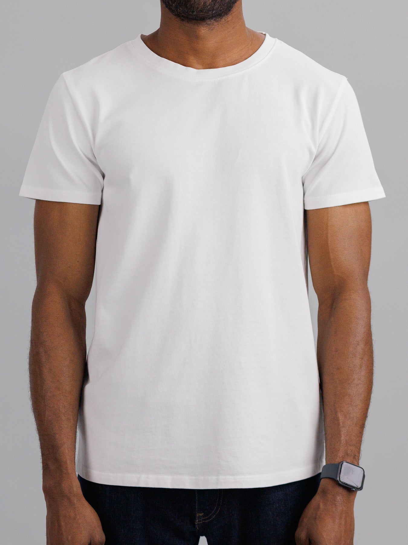 A person in a slim fit City Organic Cotton Tee by ID Menswear and dark jeans stands against a neutral background. Their face is hidden, and an armband on their left wrist adds flair to the understated look.