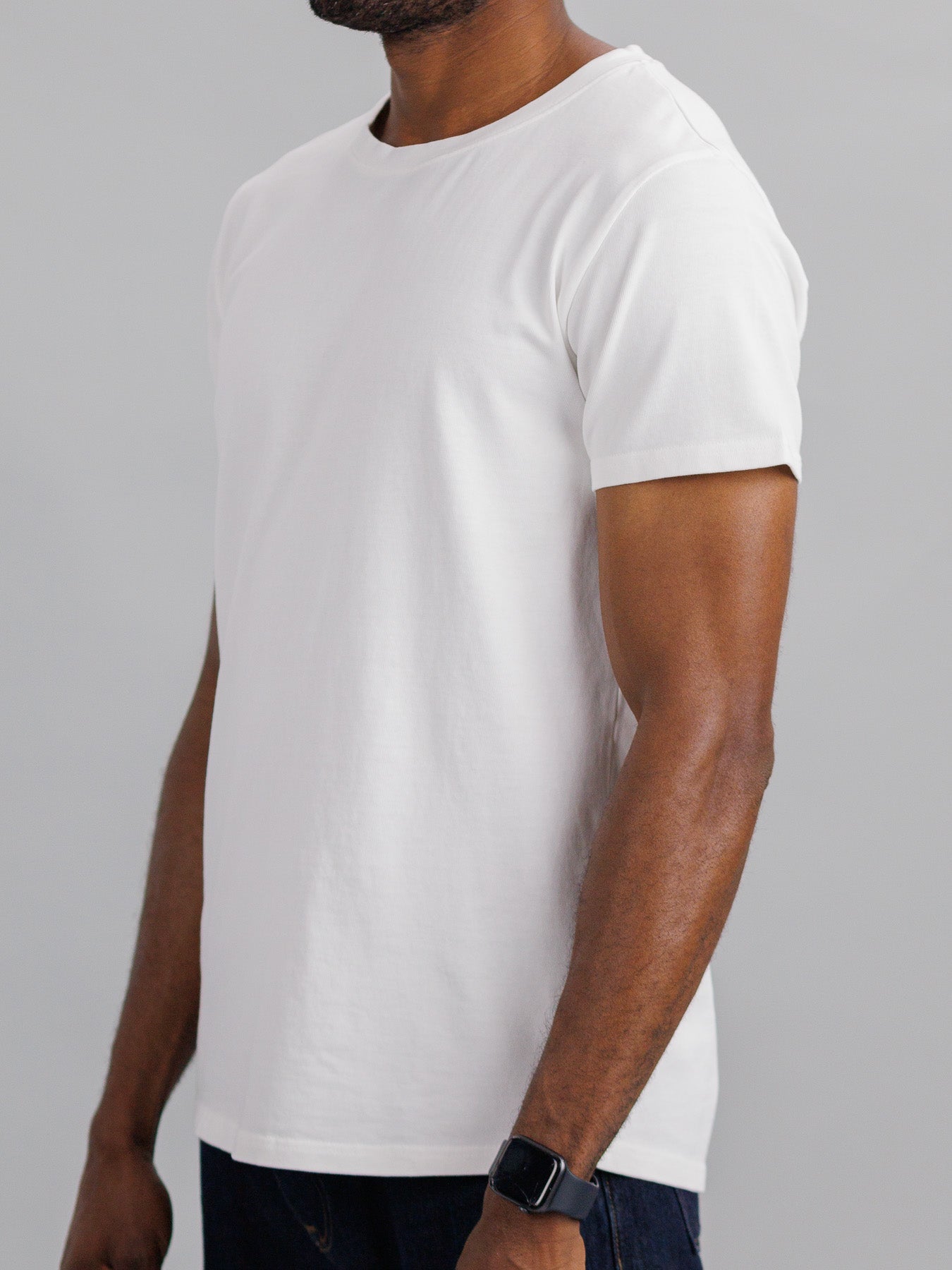 Someone wearing a slim-fit City Organic Cotton Tee by ID Menswear with dark blue jeans stands against a neutral background, showing only their upper body. Theyre also sporting a smartwatch on their wrist.