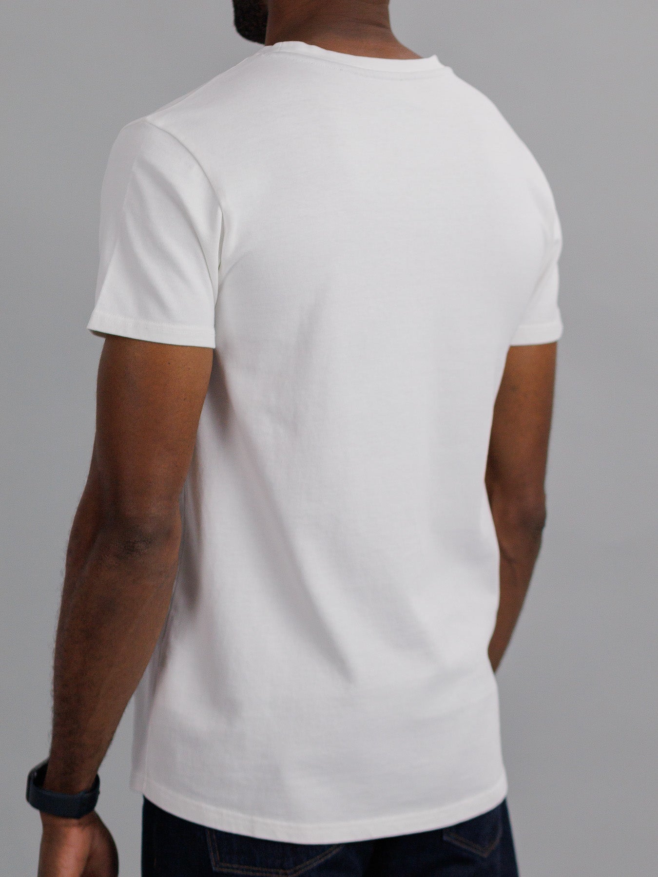 Against a gray background, someone with short hair and a black wristband on their right wrist is seen from the back in a slim fit, simple City Organic Cotton Tee by ID Menswear. The white T-shirt, made from organic cotton, pairs well with dark pants.
