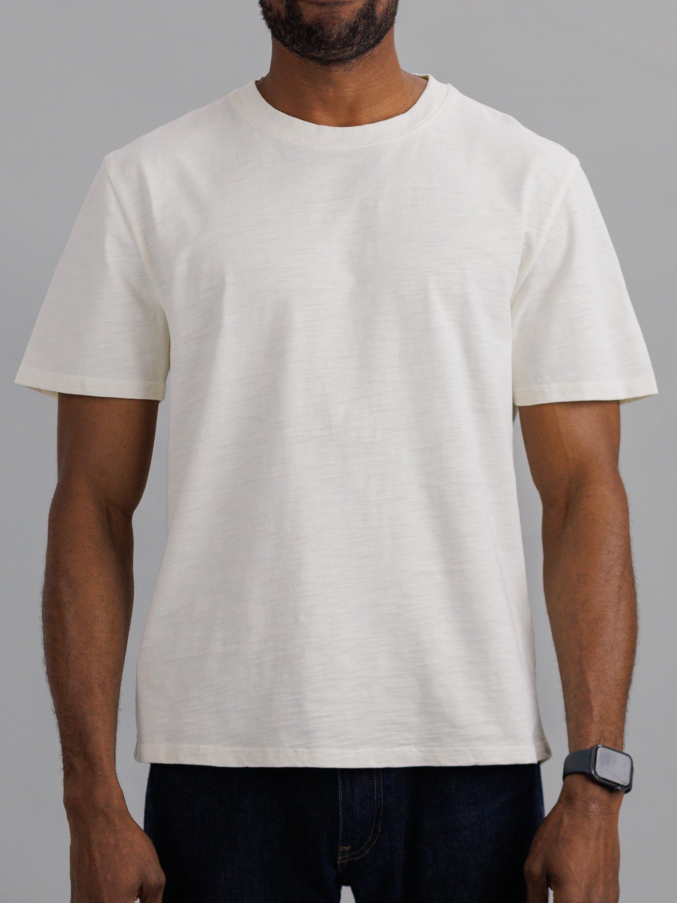 A person sports an ID Menswear Big Flip Slub Yarn Cotton Tee in plain white against a gray background. The short-sleeve, round neck tee pairs with a smartwatch on their left wrist and partially visible black pants, completing a casual yet refined style.