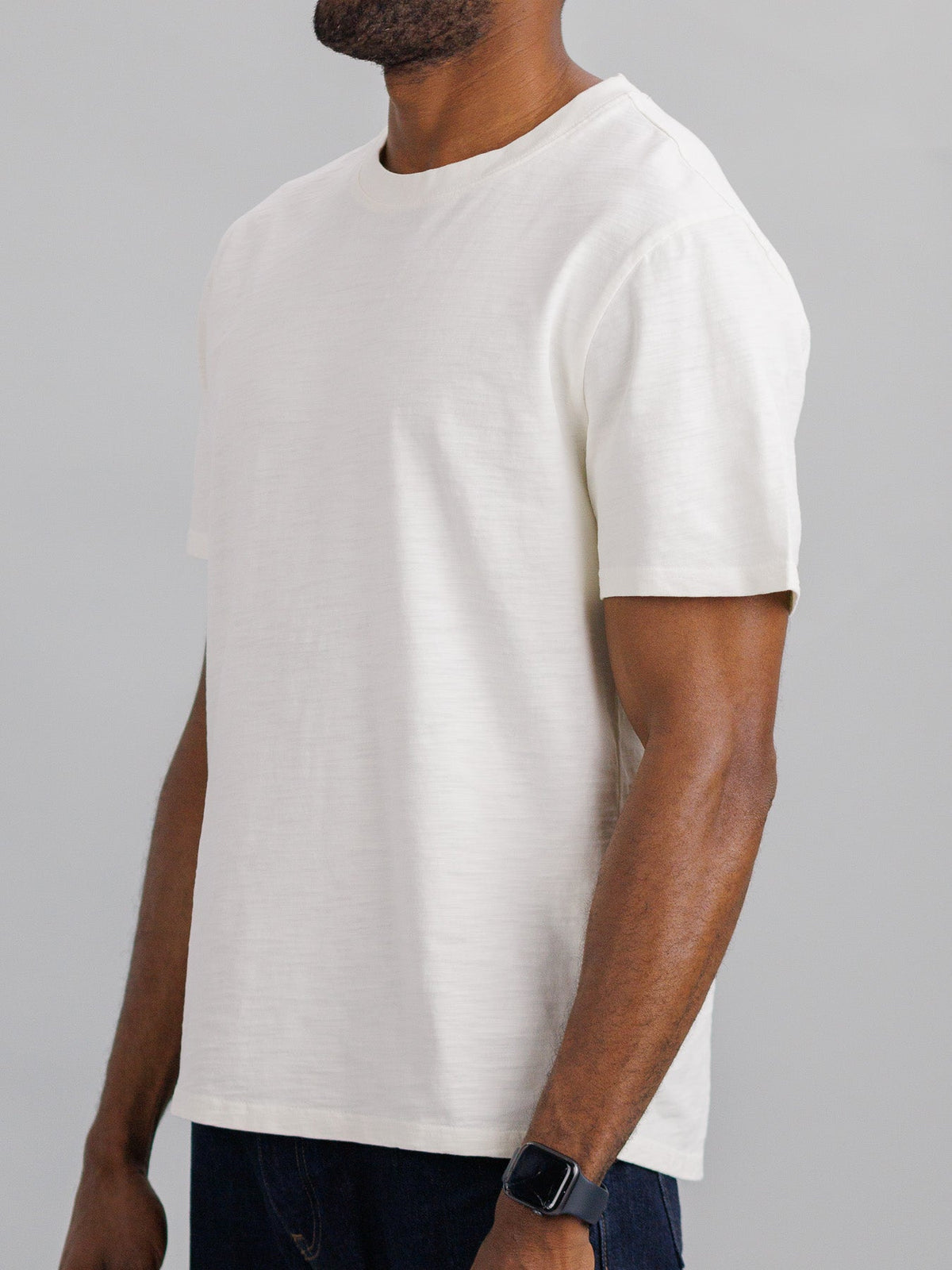 A person in ID Menswears Big Flip Slub Yarn Cotton Tee, a plain, boxy fit T-shirt made from 100% cotton, and dark jeans stands against a light gray background. Their bent arm highlights a dark wristwatch.