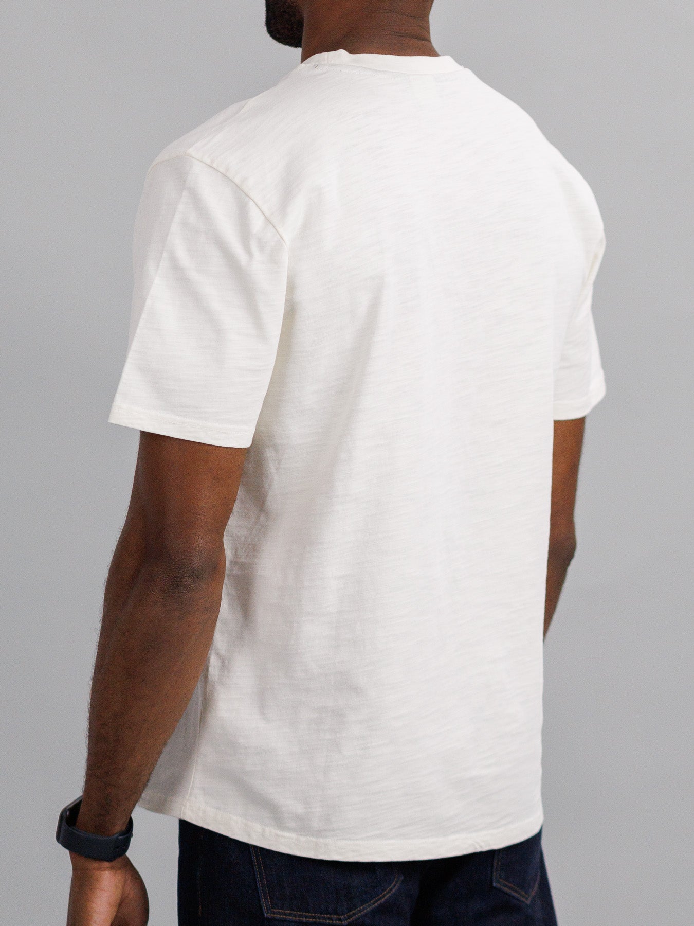 A person in a Big Flip Slub Yarn Cotton Tee by ID Menswear and dark jeans faces away from the camera. Their visible right arm sports a black smartwatch, while the tees texture adds interest against the light gray backdrop.
