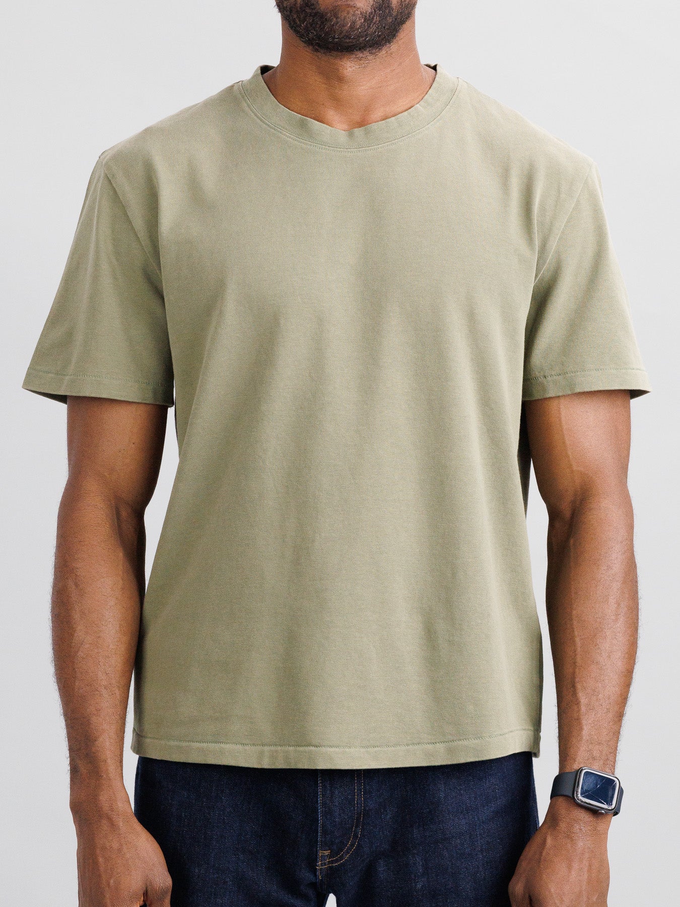 A man with a short beard and an electronic watch on his left wrist stands against a light gray background, wearing ID Menswears Freeman Organic Vintage Wash Tee in olive green and dark jeans, featuring a boxy fit with a vintage worn look.