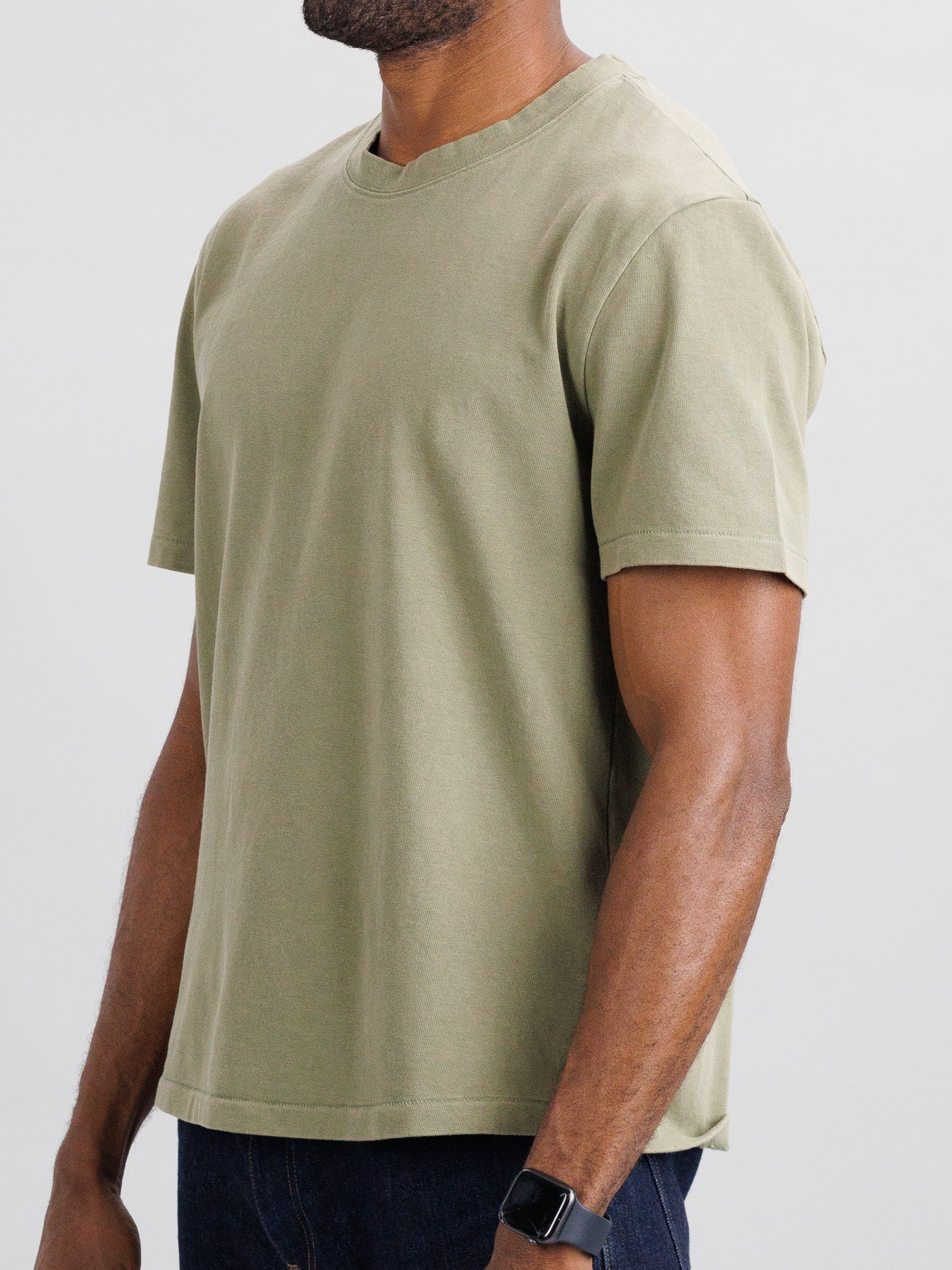 A person in a Freeman Organic Vintage Wash Tee by ID Menswear and dark jeans stands against a neutral background. The vintage olive green T-shirt complements the smartwatch on their slightly bent left wrist.