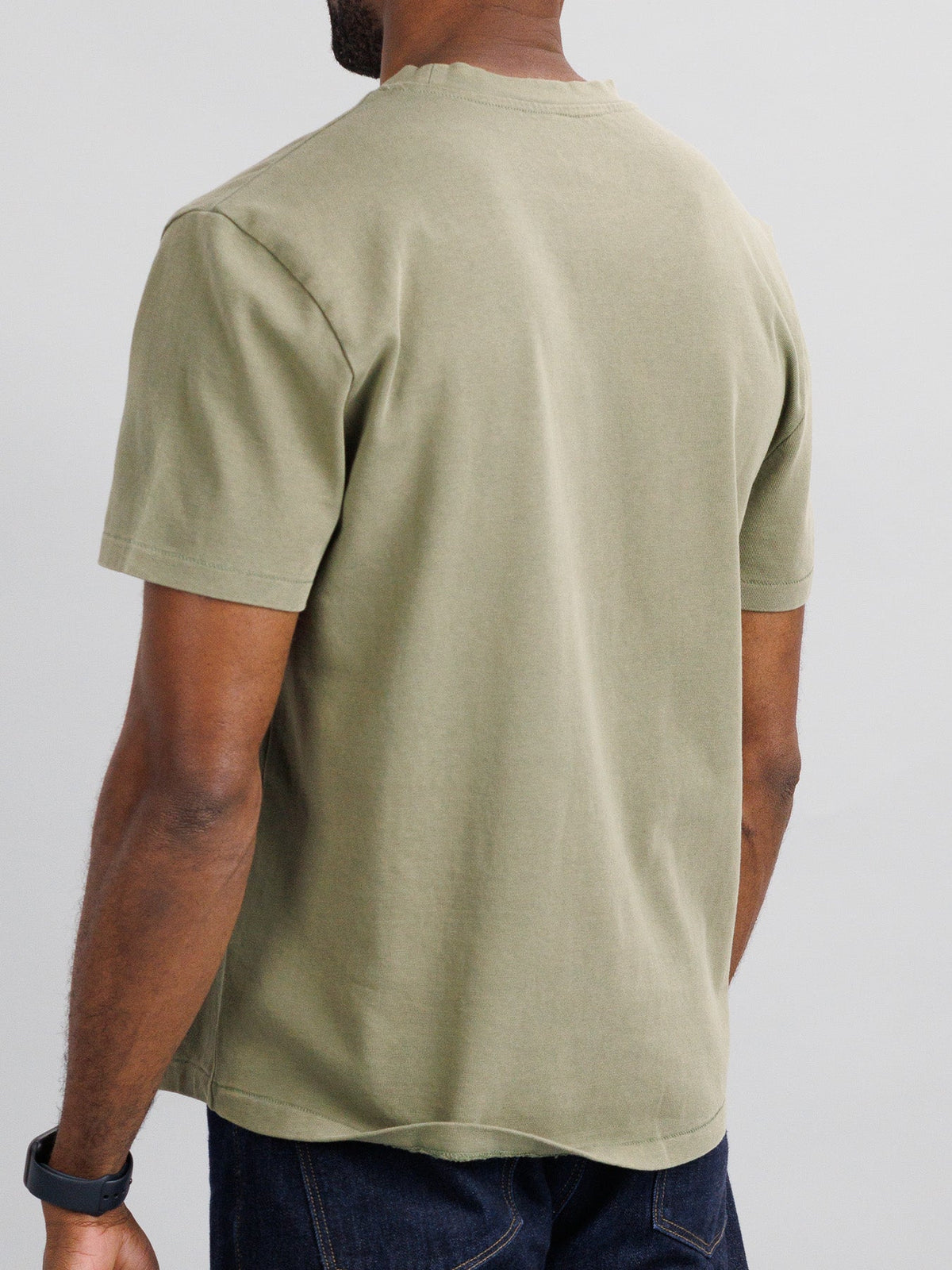 The individual in a Freeman Organic Vintage Wash Tee by ID Menswear exudes a classic vibe, pairing it with dark jeans and short hair. A black wristwatch adorns their left wrist, set against a simple light background that highlights their boxy fit look.