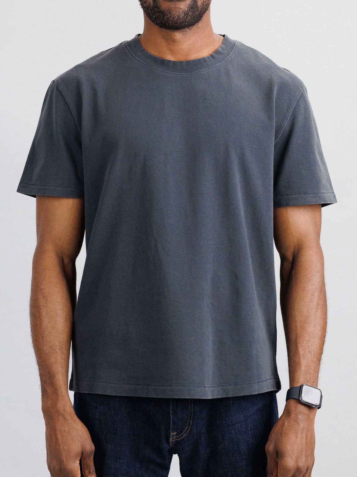 A person in an ID Menswear Freeman Organic Vintage Wash Tee, known for its plain dark gray hue and relaxed boxy fit, stands against a light gray backdrop. The shirt is paired with dark jeans, and a smartwatch adorns their left wrist; the image captures their upper body with arms at their sides.