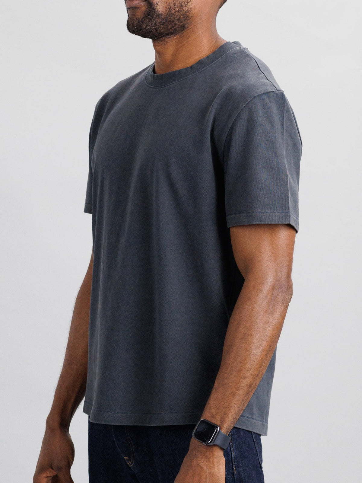 Wearing a Freeman Organic Vintage Wash Tee in dark gray and a black wristwatch, an individual stands against a light gray background. The tee by ID Menswear features a plain design with a boxy fit and crew neck, and their hands rest relaxed at their sides.