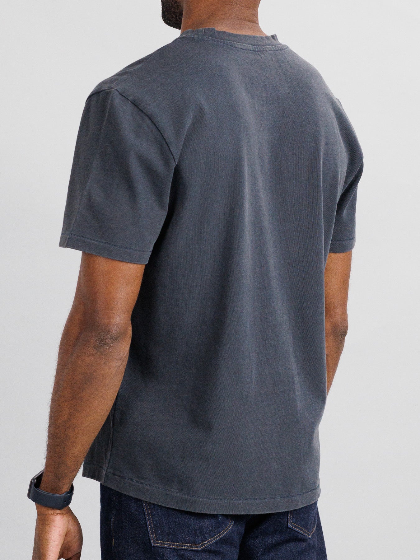 A person wearing the Freeman Organic Vintage Wash Tee in dark gray and dark jeans stands with their back to the camera. The boxy fit from ID Menswear complements short hair and a wristwatch on their left wrist against a plain, light-colored background.