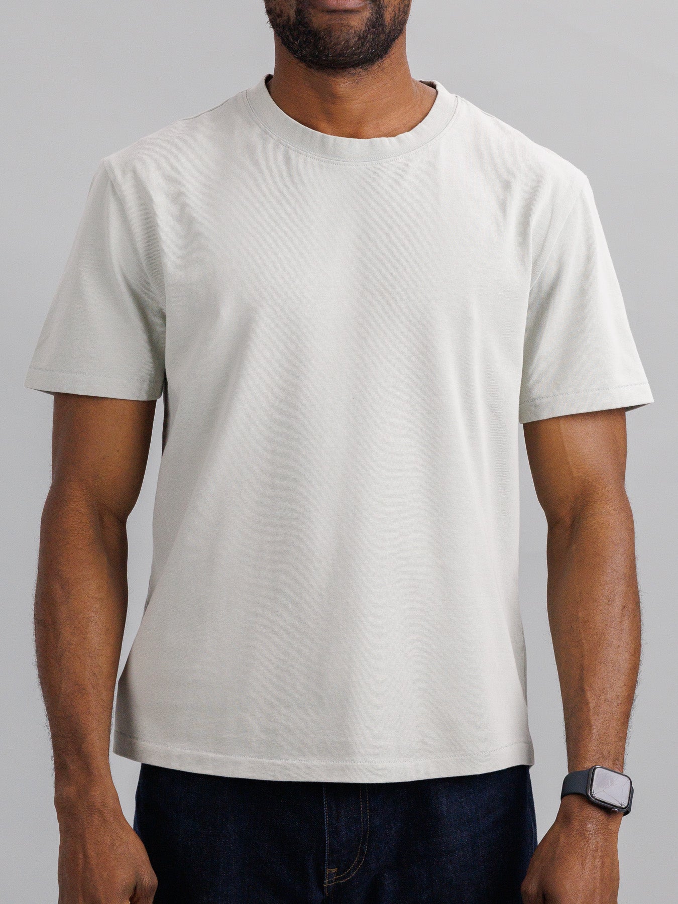 A person wearing the Freeman Organic Vintage Wash Tee by ID Menswear and a smartwatch stands with relaxed arms. The plain light gray tee has a vintage worn look, complemented by the soft, neutral background.