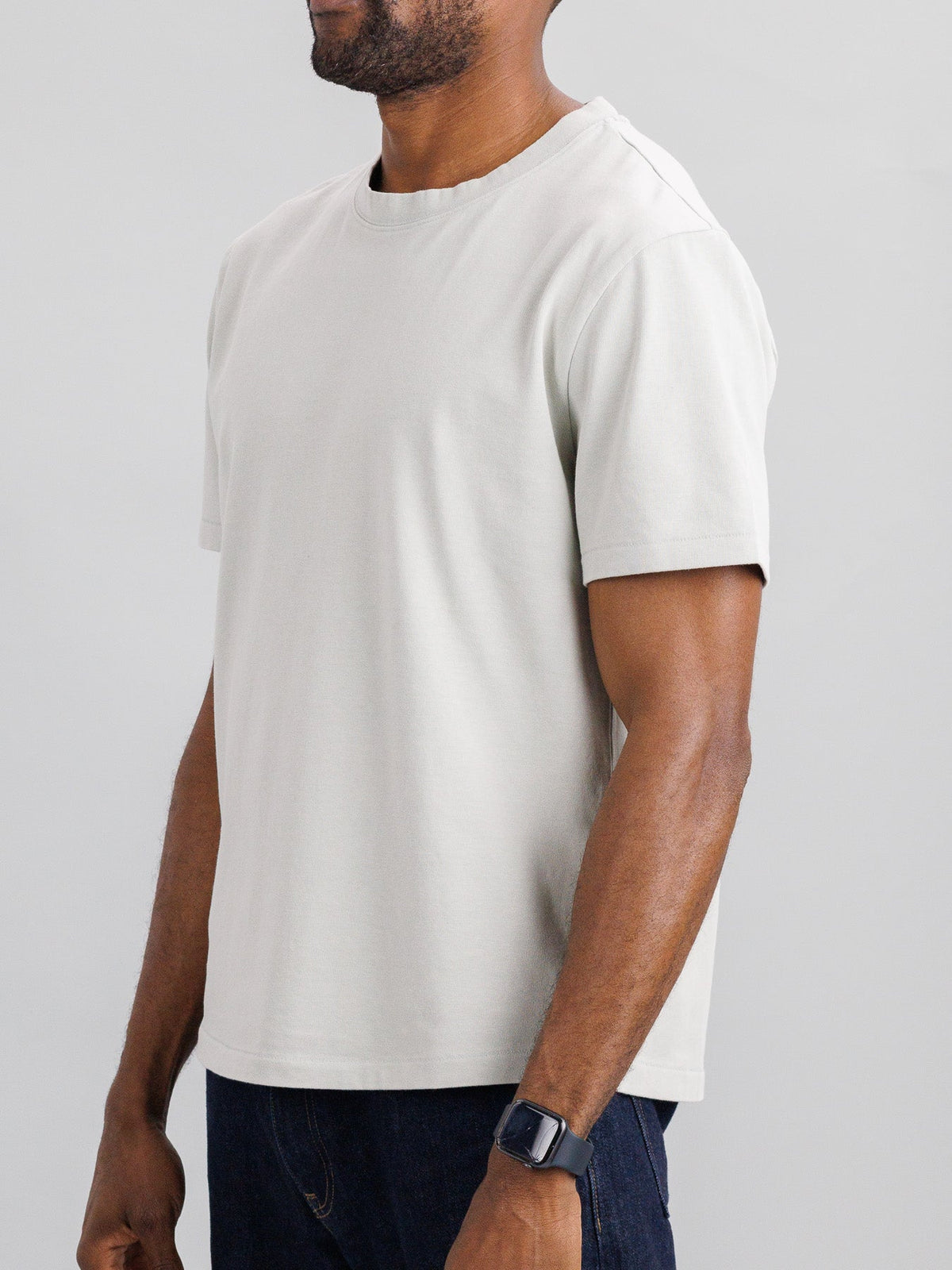 A person in an ID Menswear Freeman Organic Vintage Wash Tee, featuring a boxy fit and light gray color, with a smartwatch on their wrist, stands against a plain background.