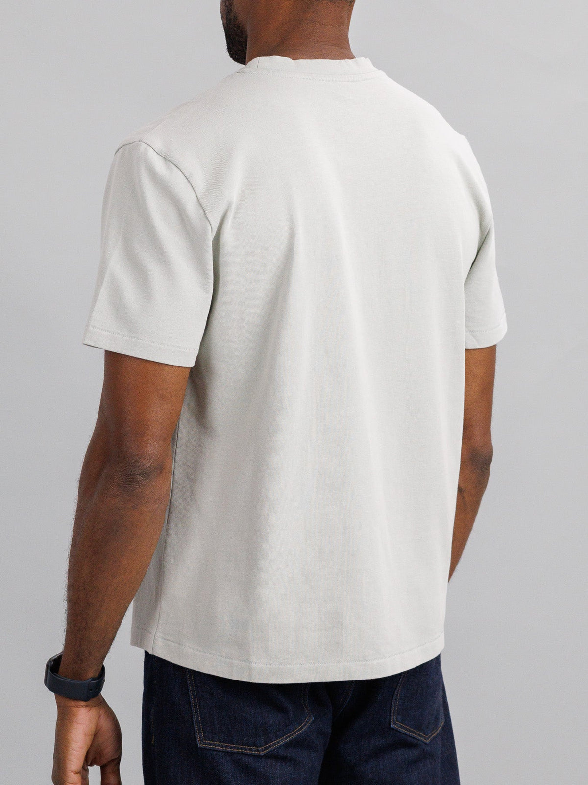 A person in the ID Menswear Freeman Organic Vintage Wash Tee and dark jeans stands, showcasing the shirts boxy fit. Their left arm displays a black wristwatch against a plain gray background that enhances the tees subtle vintage look.