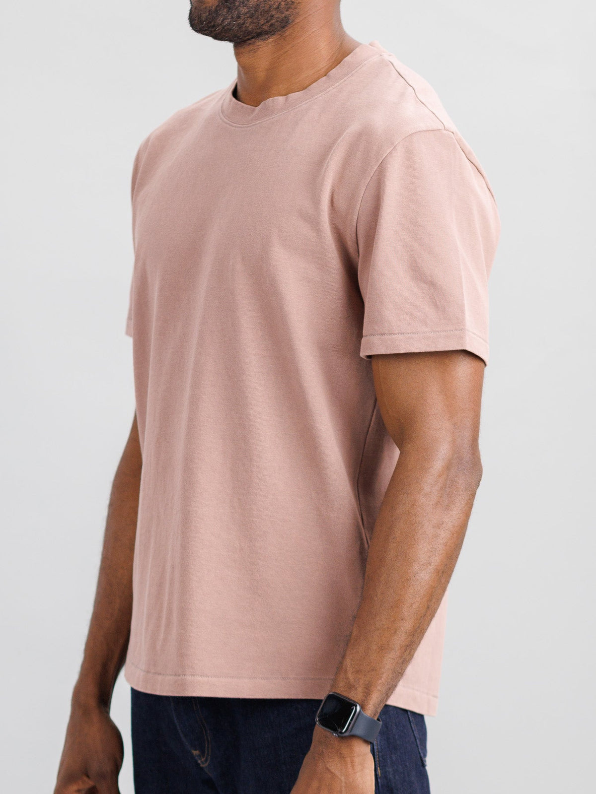 A person dressed in ID Menswears Freeman Organic Vintage Wash Tee, characterized by its plain light brown hue and vintage worn appeal, pairs it with dark jeans and a black smartwatch against a light gray backdrop. The shirts boxy fit is clear despite the headless image.