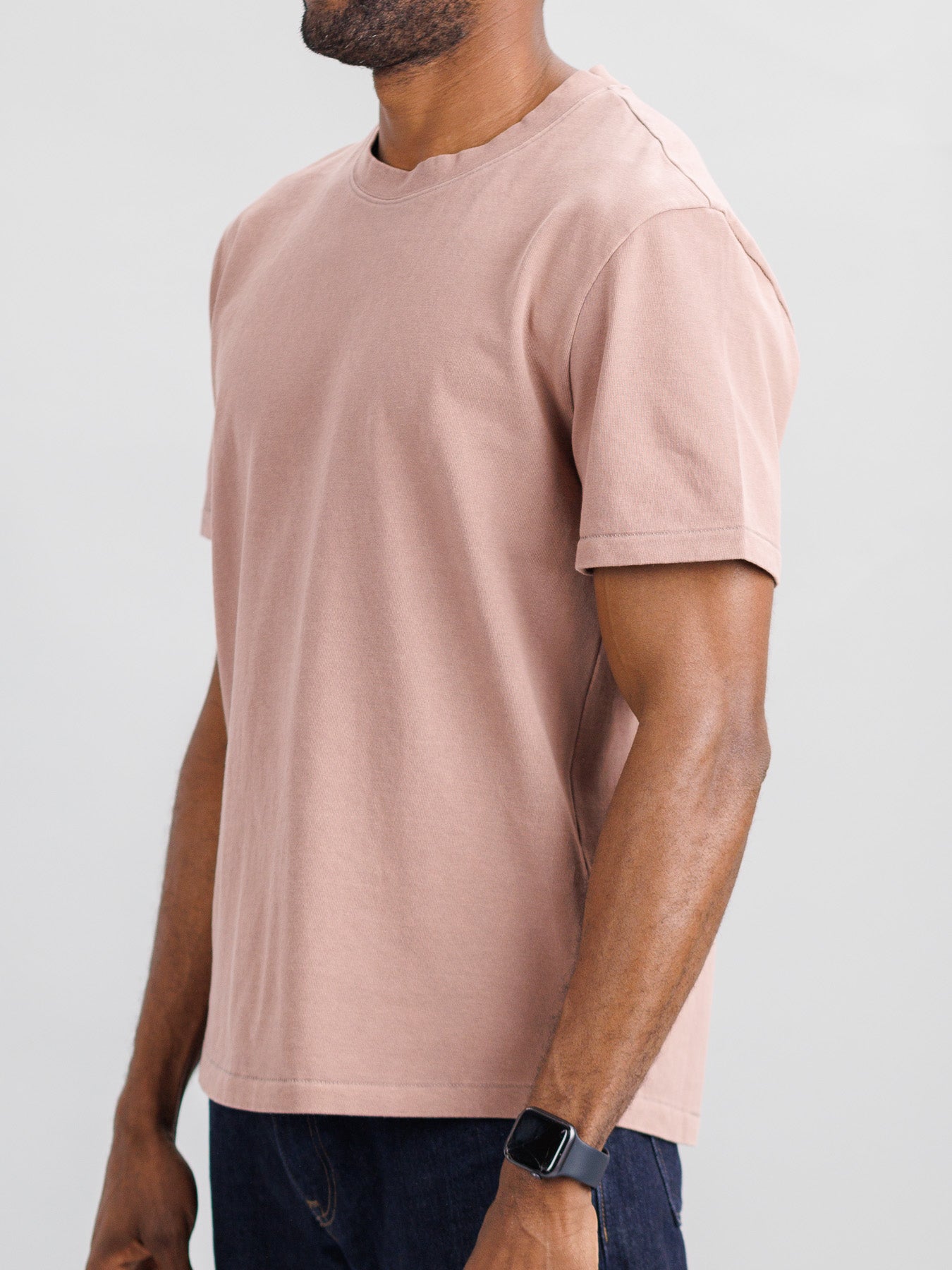 A person dressed in ID Menswears Freeman Organic Vintage Wash Tee, characterized by its plain light brown hue and vintage worn appeal, pairs it with dark jeans and a black smartwatch against a light gray backdrop. The shirts boxy fit is clear despite the headless image.