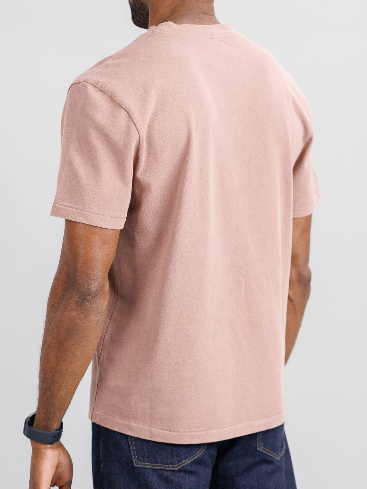 A person wearing an ID Menswear Freeman Organic Vintage Wash Tee in light pink and dark jeans is facing away from the camera, with a smartwatch on their left wrist against a neutral backdrop.