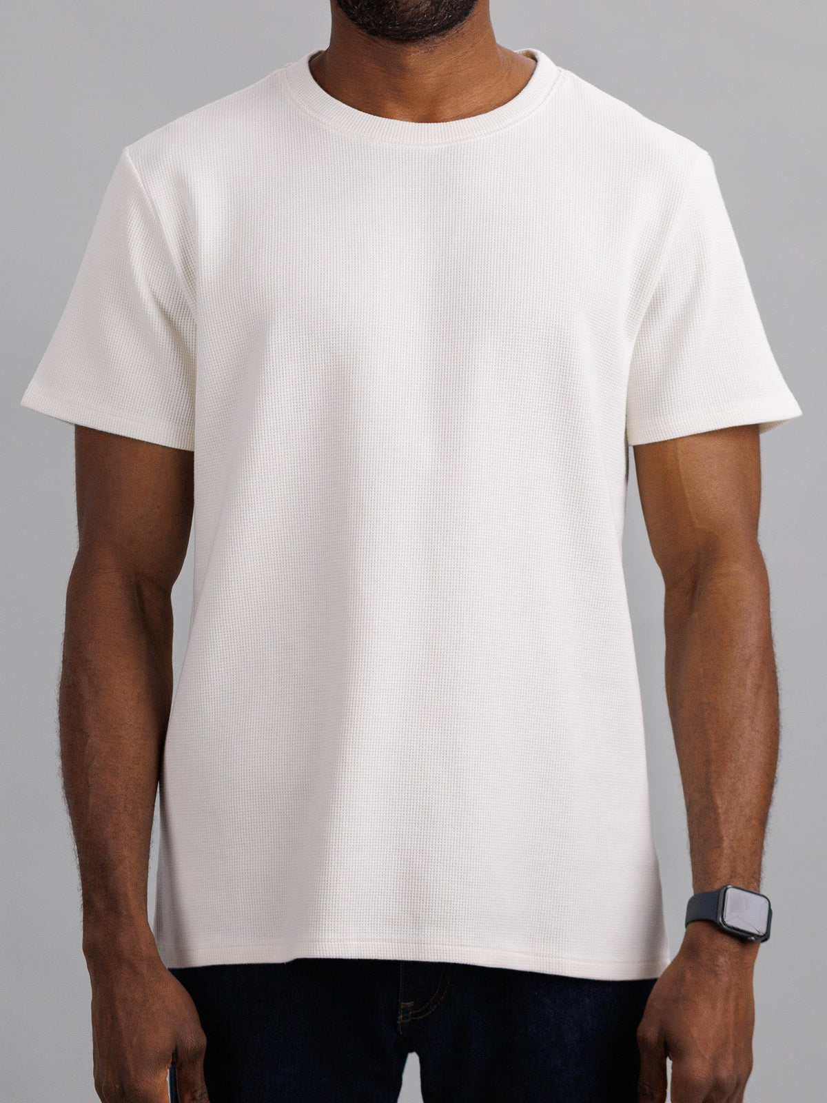 Dressed in a casual ID Menswear Urban Waffle Knit Tee with dark pants, an individual stands against a neutral backdrop, showcasing a smartwatch on their visible left wrist.